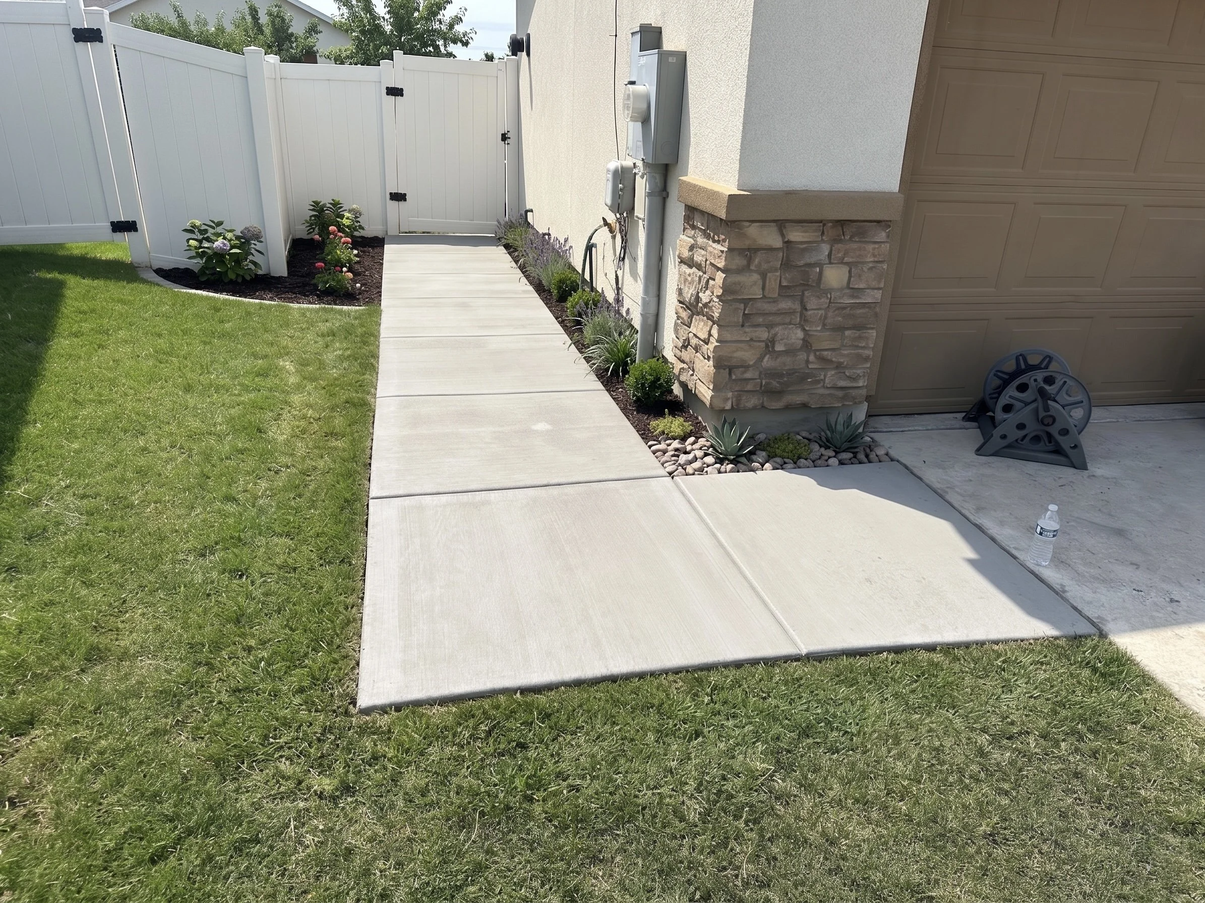A backyard with a lawn, concrete sidewalk, white fence and gate, garden bed with flowers, siding of a house, a stone pillar, and a section of an attached garage door. A small water bottle and wheel parts are on the ground near the garage.
