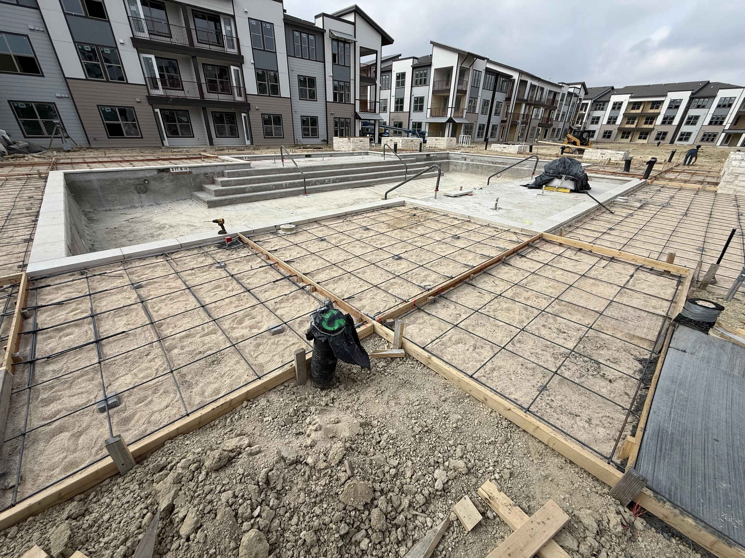 Construction site of a community pool area with steps, surrounded by framing and rebar for concrete, in front of modern apartment buildings under cloudy sky.