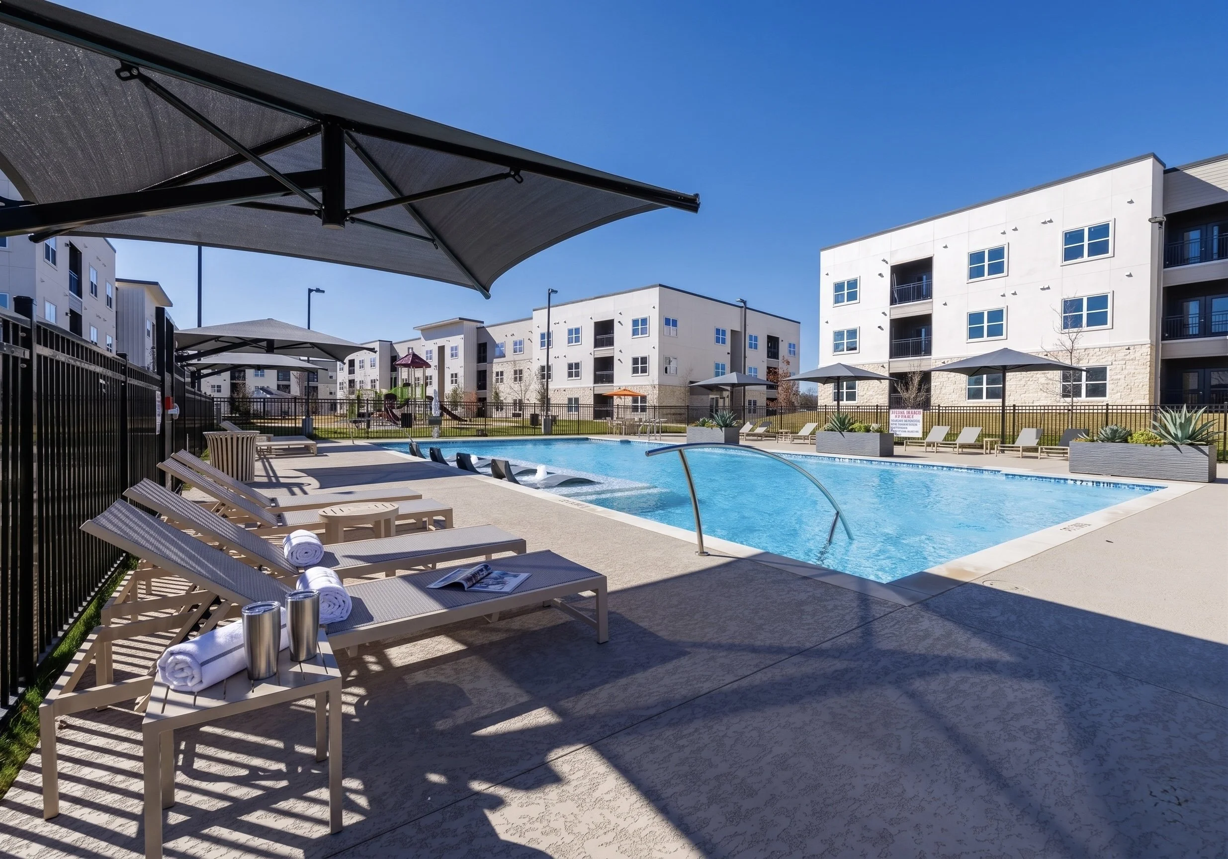 A residential outdoor swimming pool area with lounge chairs, umbrellas, and a playground in the background, surrounded by a black metal fence and modern apartment buildings.