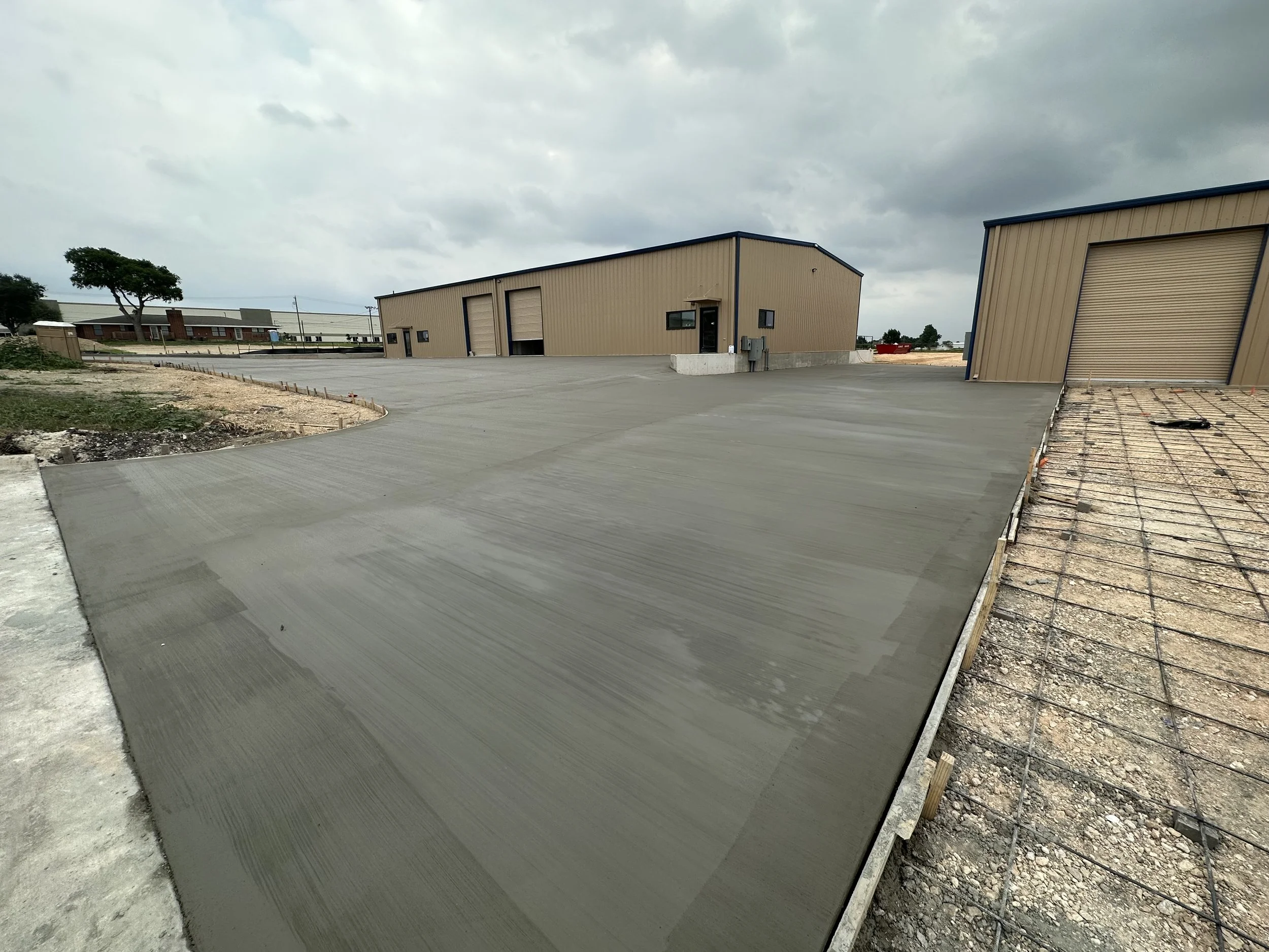 A newly poured concrete slab in front of two beige industrial metal buildings under a cloudy sky, with a partially completed section on the right showing rebar and formwork for additional concrete.