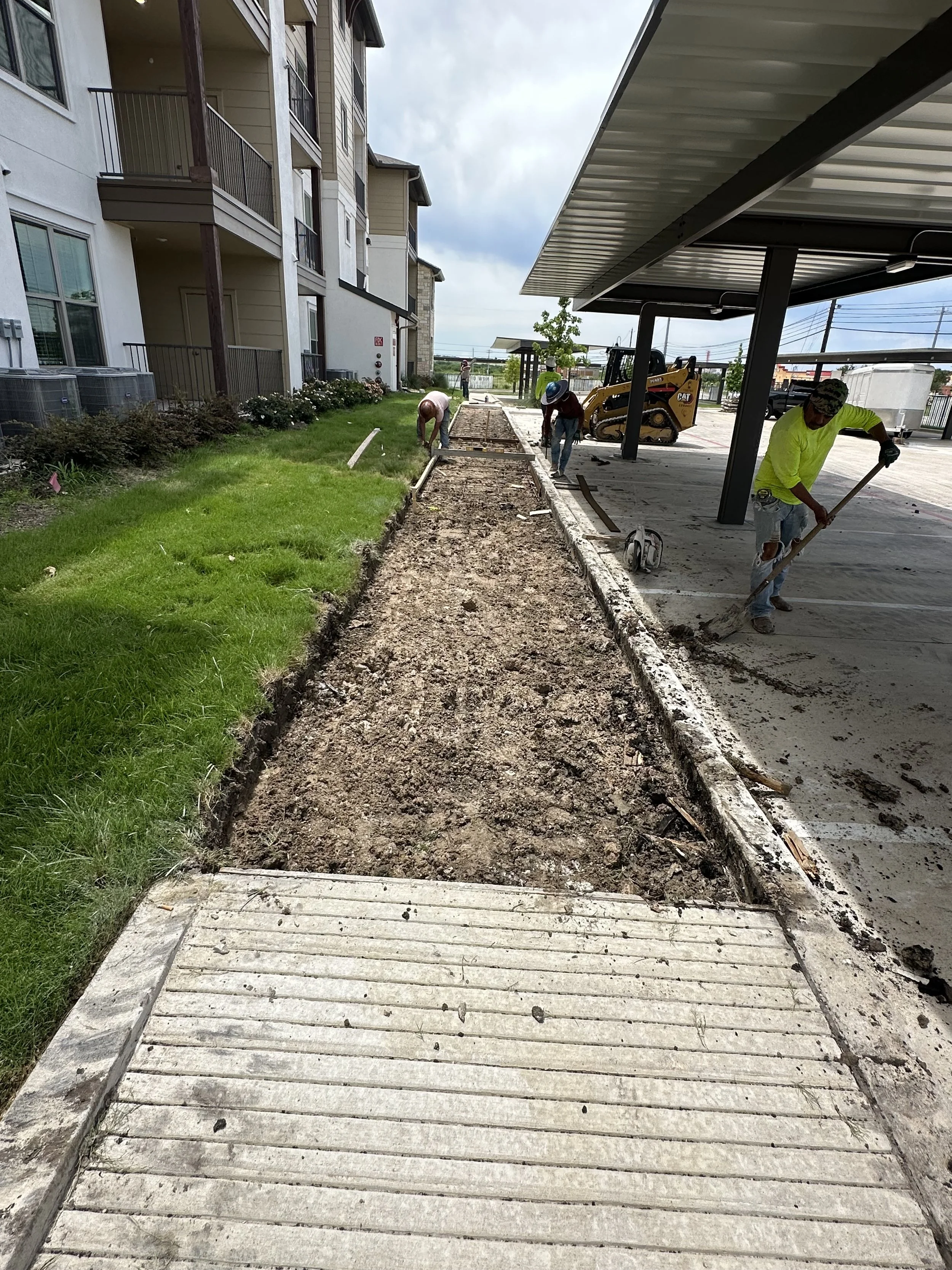 Construction workers working on a sidewalk section beside a parking lot, with an apartment building on the left and a covered parking area on the right, under cloudy skies.