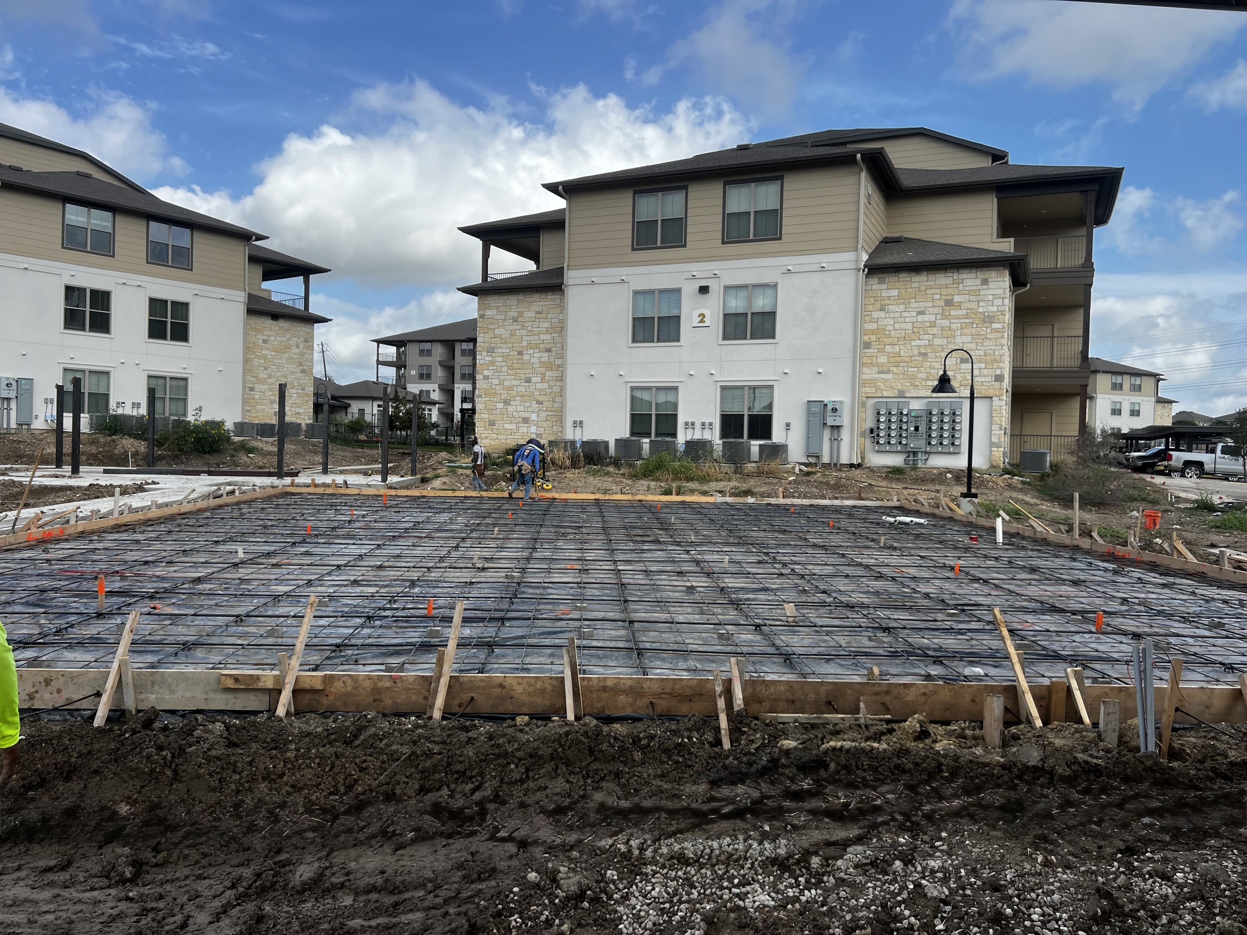 Construction site with rebar for concrete foundation in front of multi-story residential buildings with beige siding and stone accents under a cloudy sky.