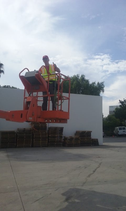 A construction worker standing on an orange cherry picker lift, giving a thumbs-up gesture, wearing a safety vest and cap, with stacked pallets and a white wall in the background.
