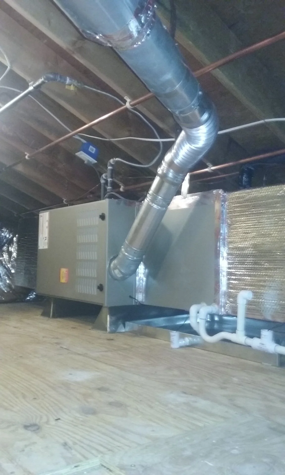 Mechanical HVAC unit with metal ductwork and pipes on a wooden floor in an unfinished basement.