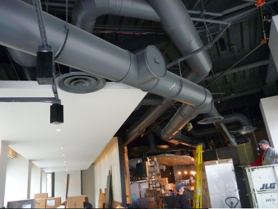 Construction workers in an indoor building under renovation, with exposed ceiling pipes and ductwork, and tools and equipment around.
