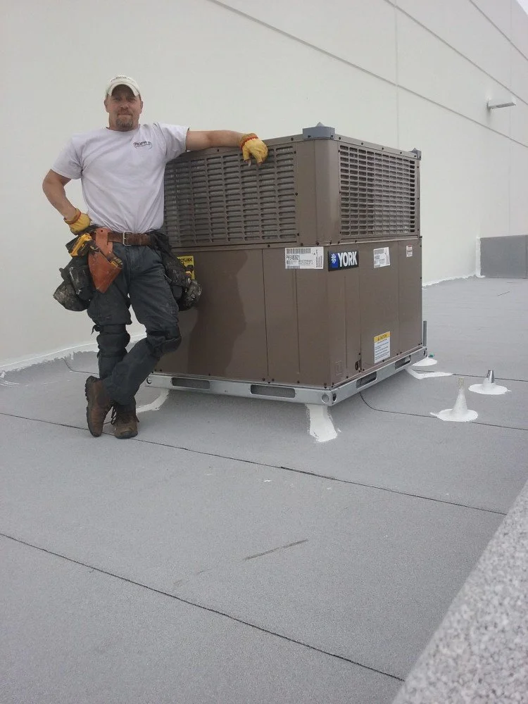 A male technician in work attire with tools attached to his belt, standing next to an industrial air conditioning or HVAC unit on a flat rooftop.