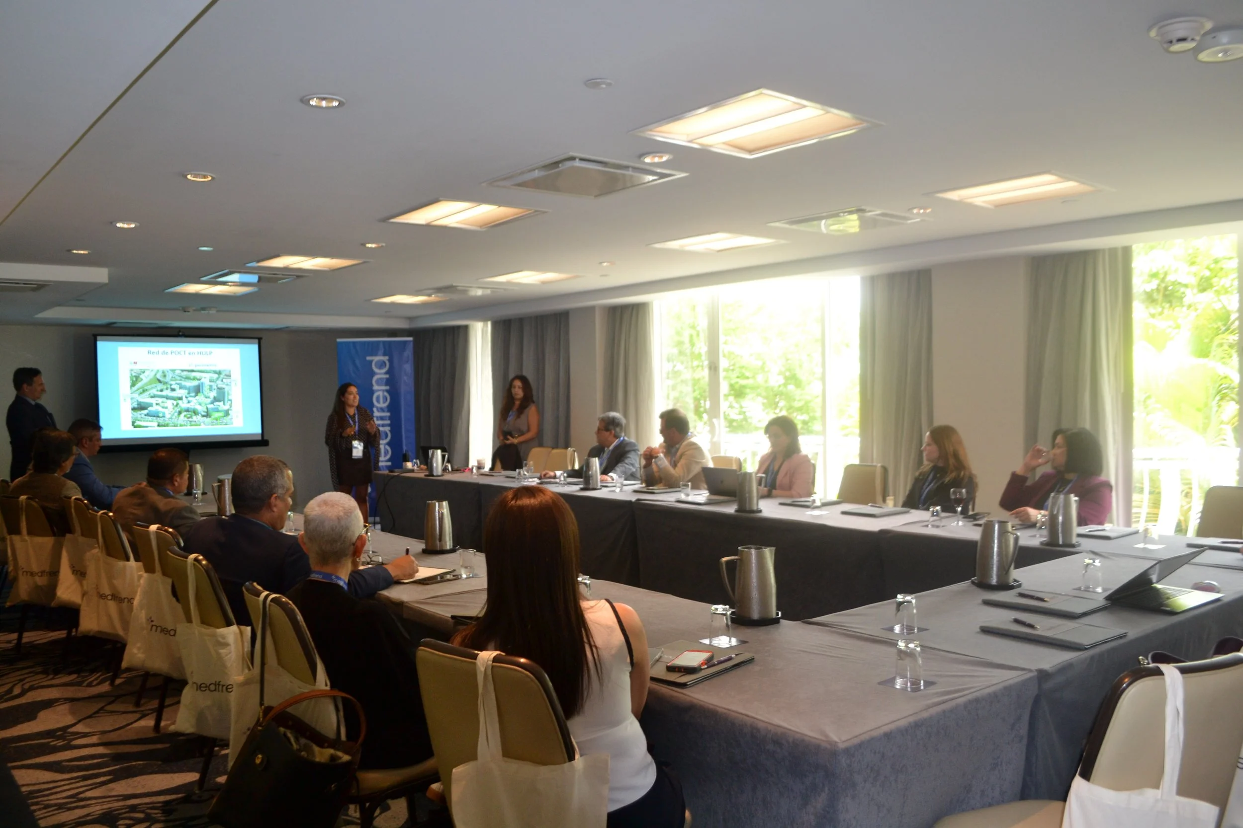 Business conference room with attendees seated around a U-shaped table, a presentation being shown on a screen, and two women standing at the front.