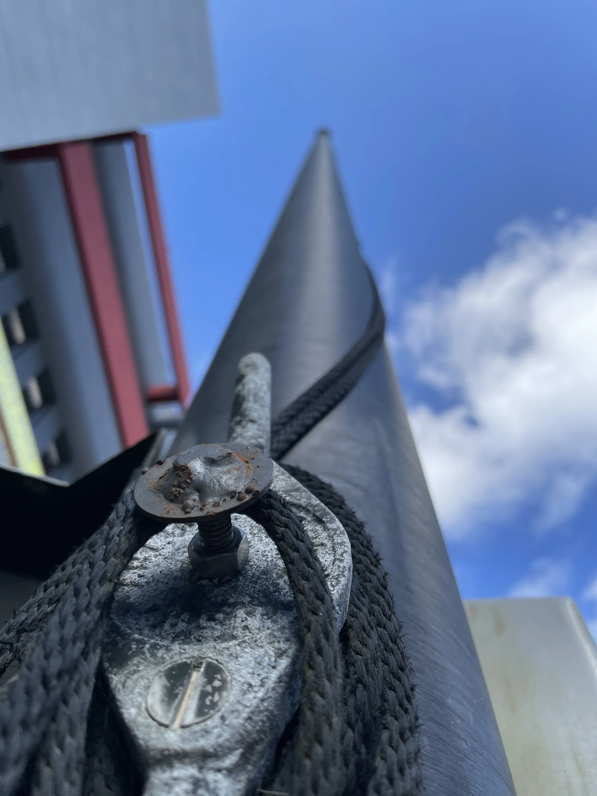 Close-up view of a metal structure with a cable secured by a metal clamp and a screw, extending vertically toward a blue sky with some clouds.