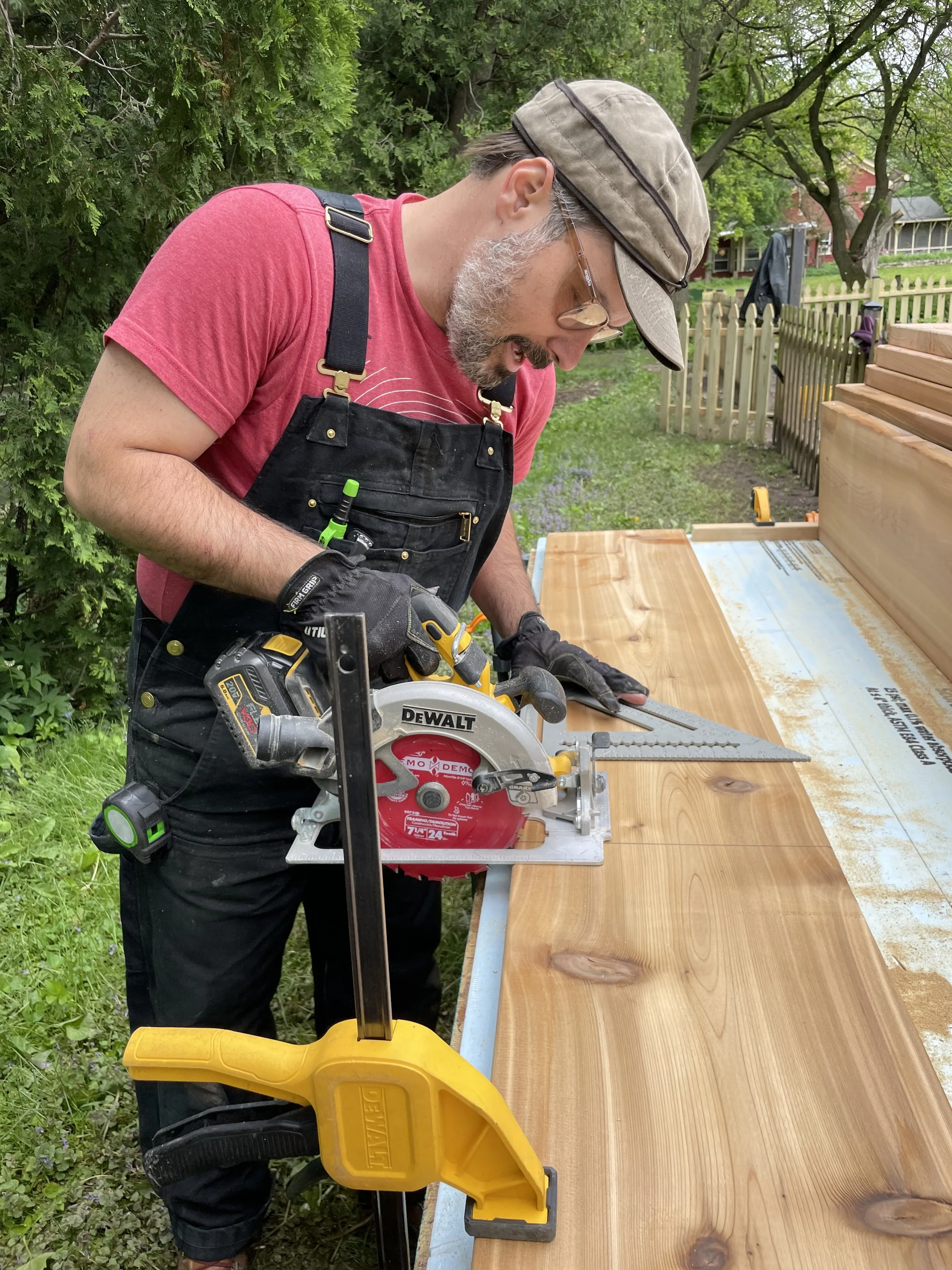 A man with a beard and glasses wearing a hat, red T-shirt, black overalls, and gloves is using a circular saw to cut wood outdoors. He is standing at a workbench with green trees and a fence in the background.