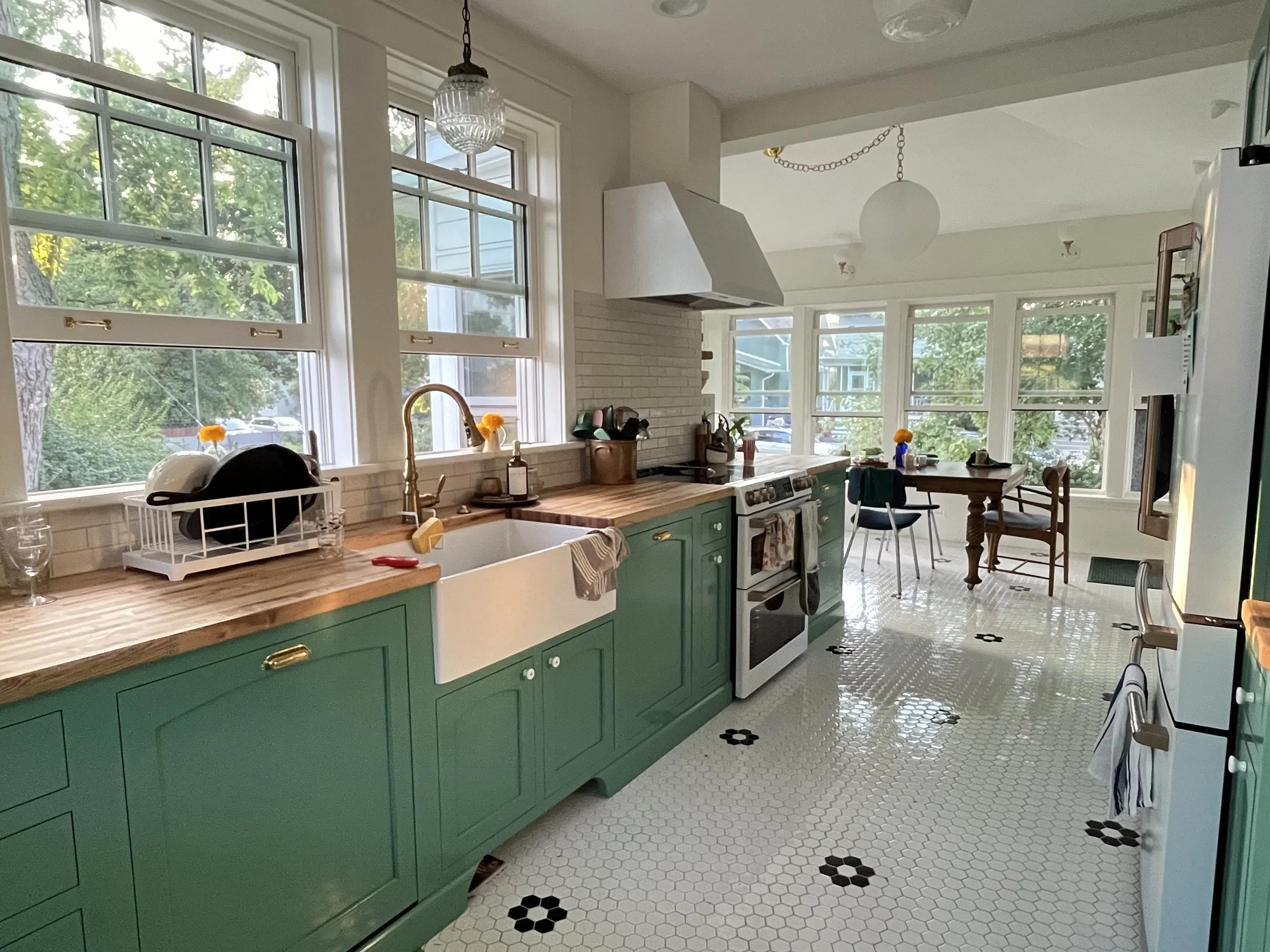 Bright kitchen with green cabinets, white farmhouse sink, wooden countertop, and large windows. There are dishes drying on a rack, a stove with a range hood, and a small dining area with a round table and chairs. The floor is white hexagonal tiles with black flower patterns.