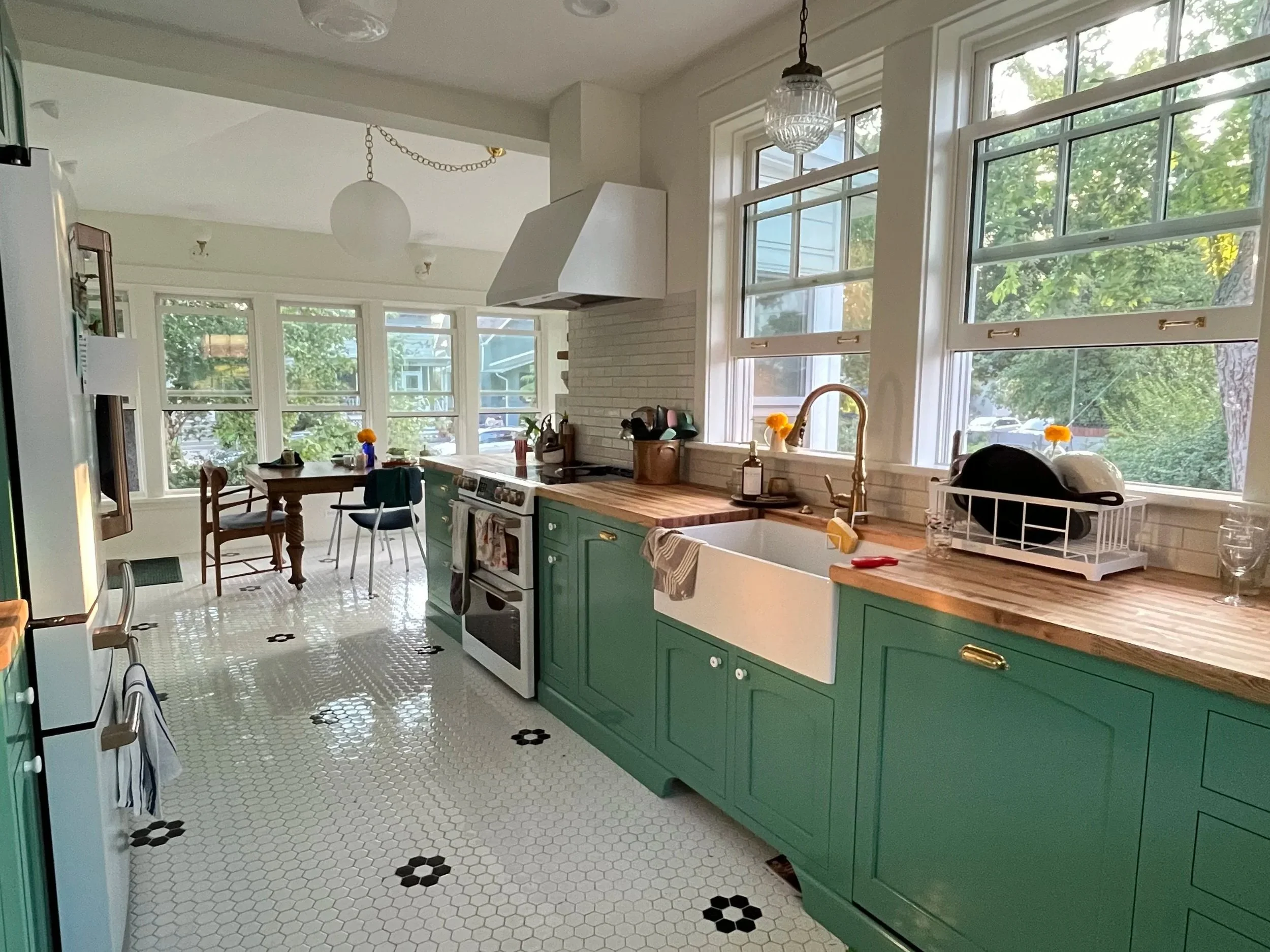 A bright kitchen with green cabinets, a white farmhouse sink, and large windows letting in natural light in Chicago. There is a wooden table with chairs in the background and a tiled floor with small black accents.