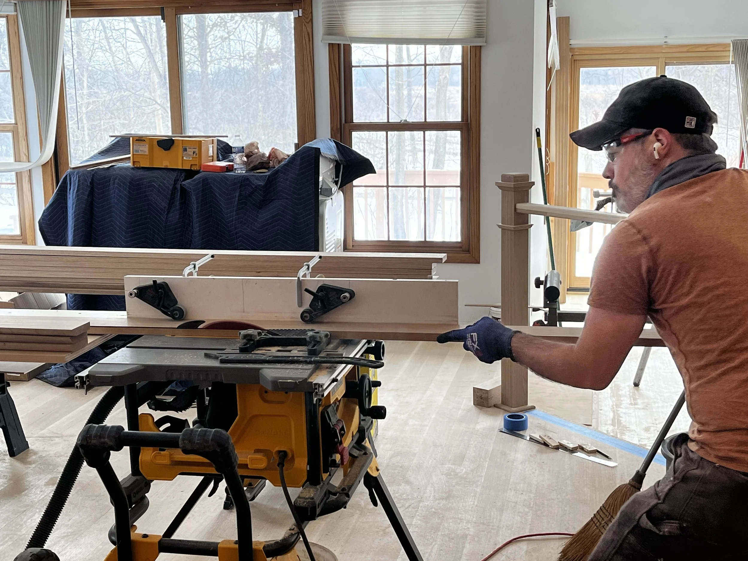 A man wearing glasses, a black cap, a brown shirt, and construction gloves is working on a woodworking project, cutting a piece of wood with a table saw inside a bright room with large windows showing trees outside.