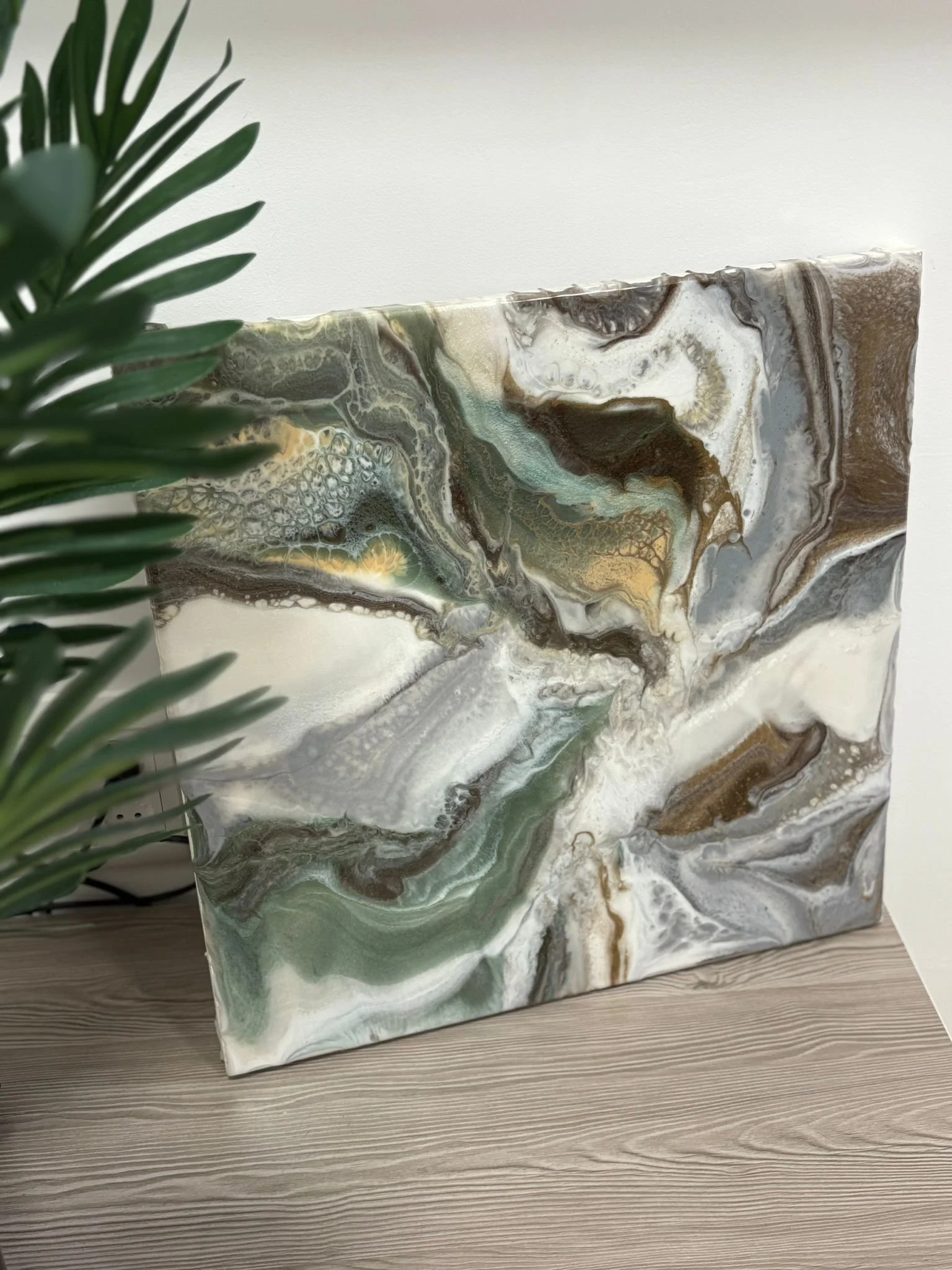 Abstract marbled painting with shades of green, white, brown, and gold leaning against a wall, partially obscured by a green plant with long, pointed leaves.