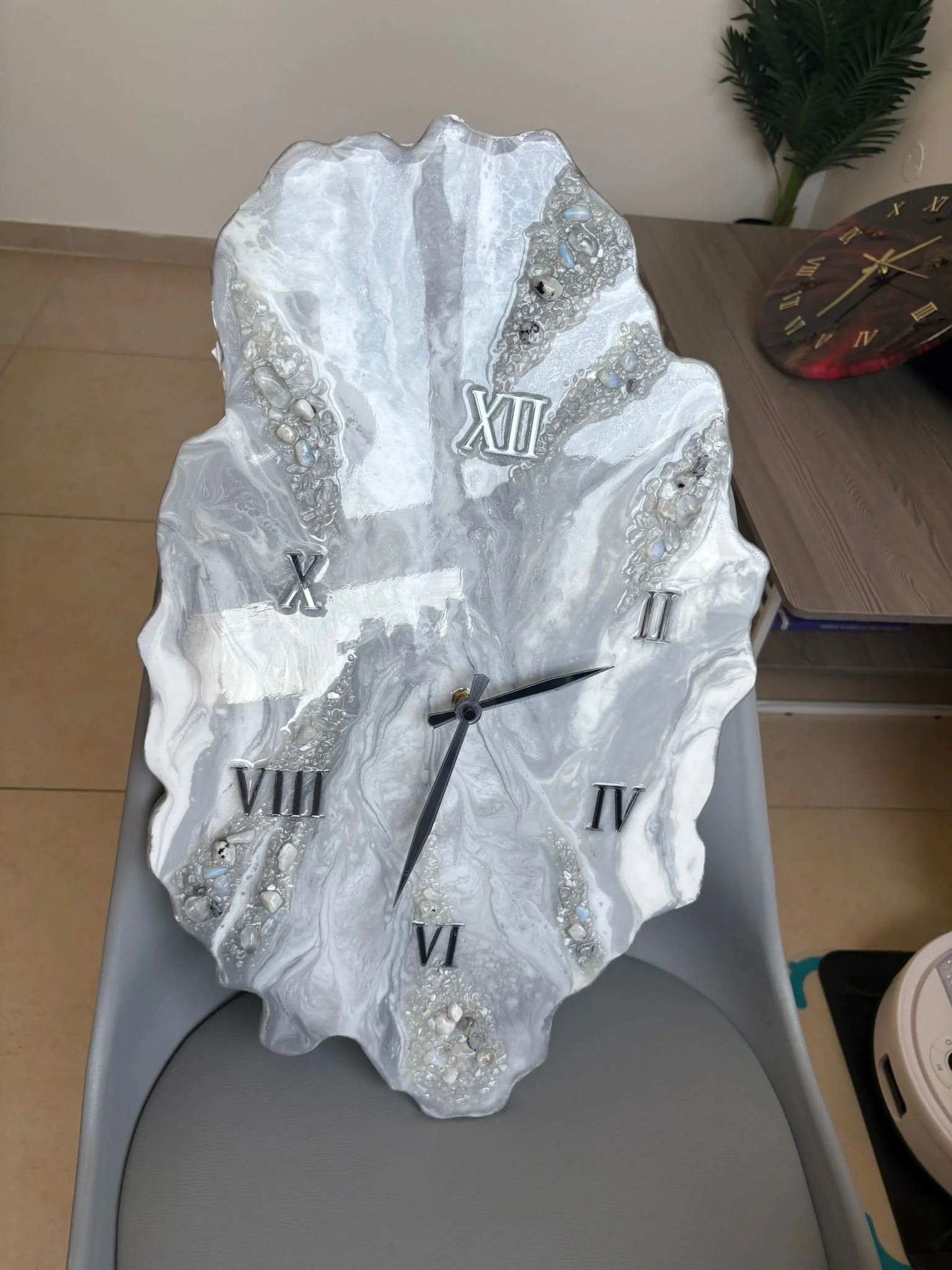 A decorative clock with a marbled, irregular-shaped face, Roman numerals, and black clock hands, placed on a gray surface.