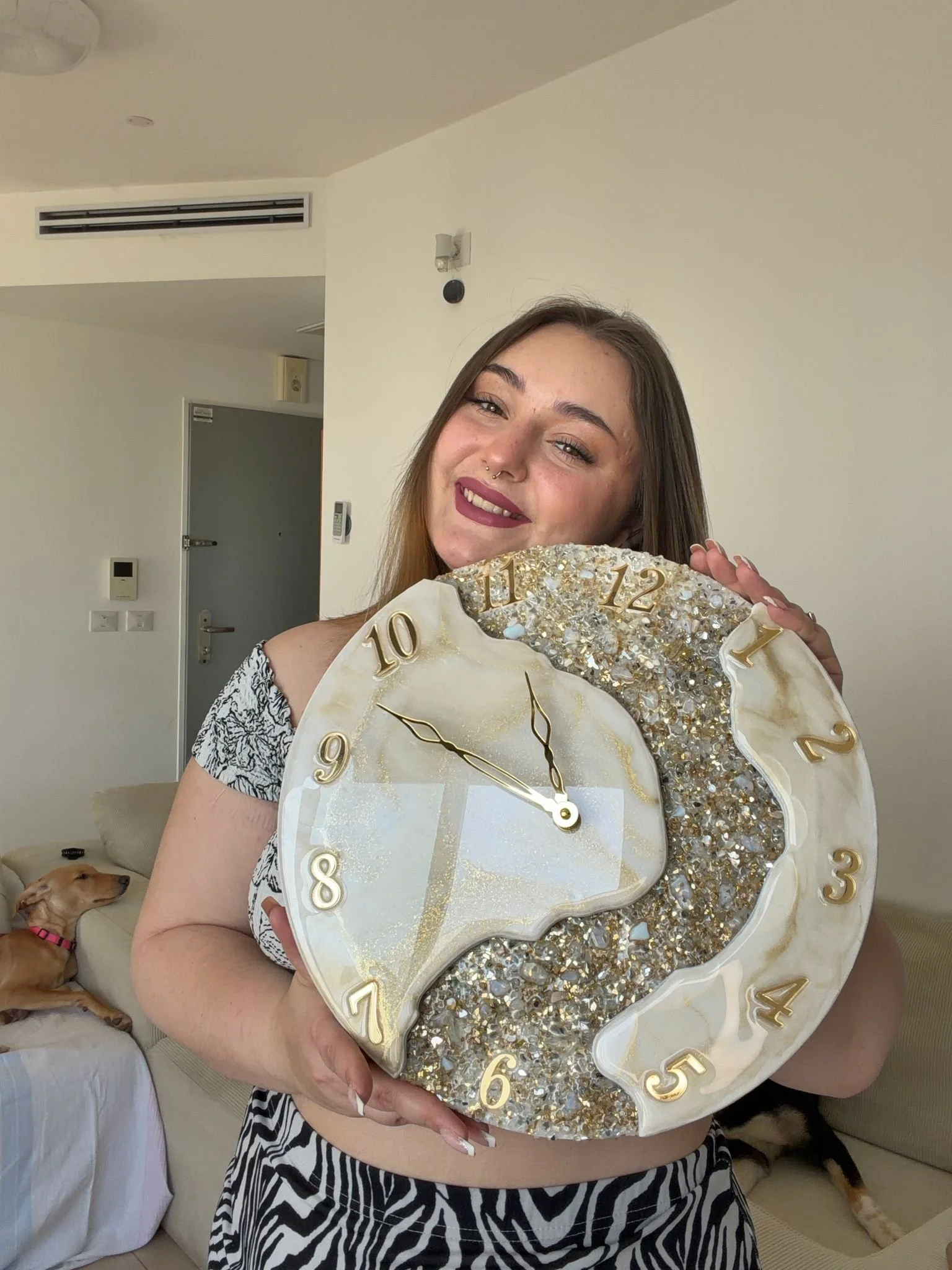 A smiling woman with long hair holding a decorative clock with gold accents and a glittery design, standing in a room with a dog lying on a sofa in the background.