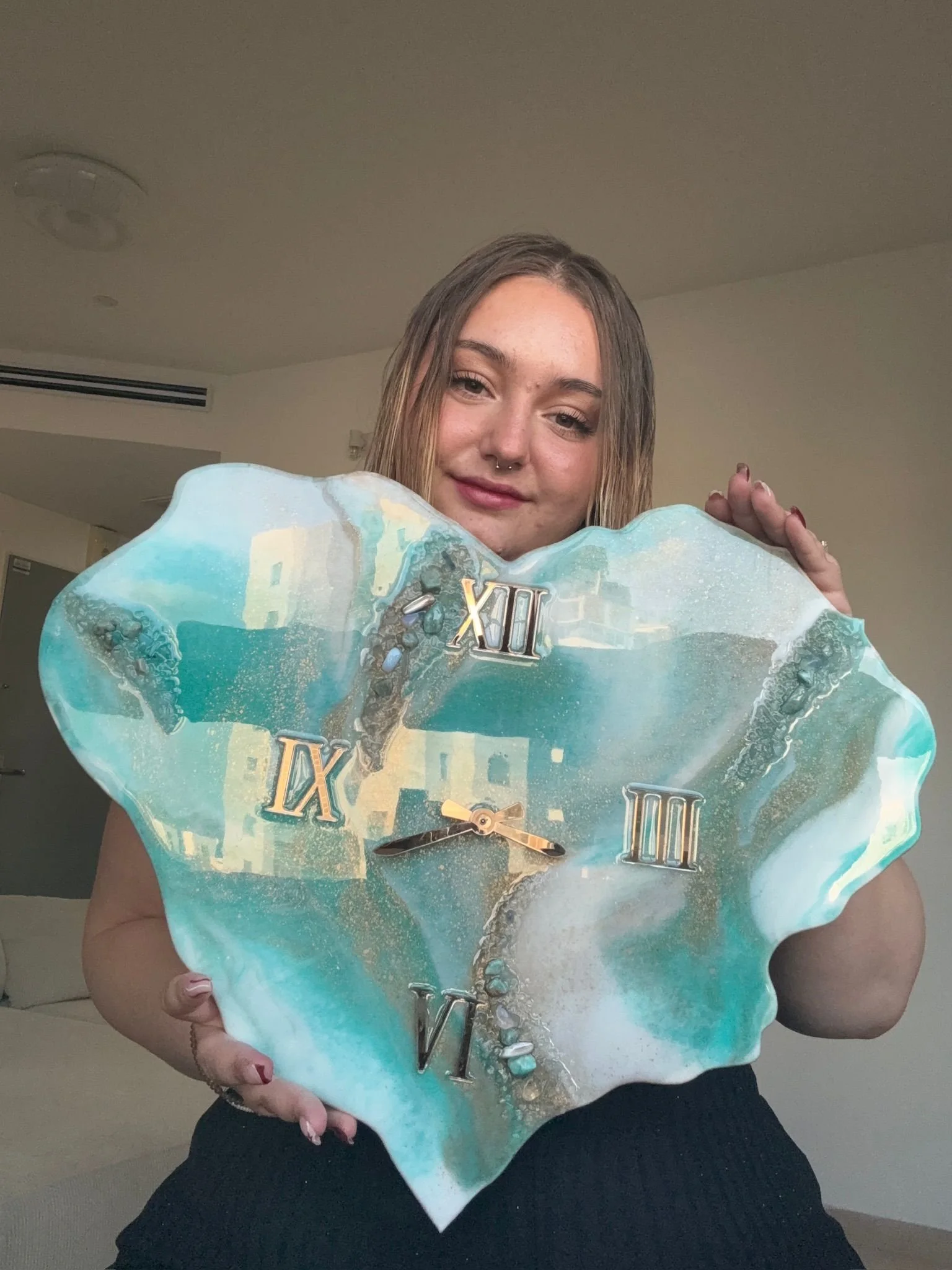 A young woman with shoulder-length hair holds a large, irregularly shaped clock with a marble-like design in shades of blue, white, and beige. The clock has Roman numerals and gold-colored hands.