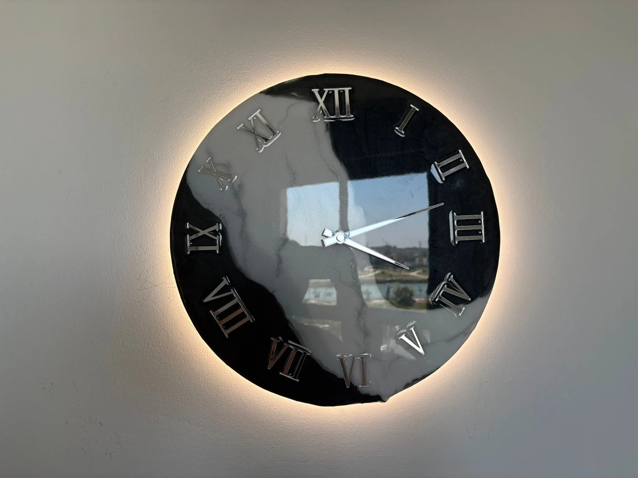 Round wall clock with black background, silver Roman numerals, silver hands, and a luminous backlight, reflecting a window and an outdoor scene.