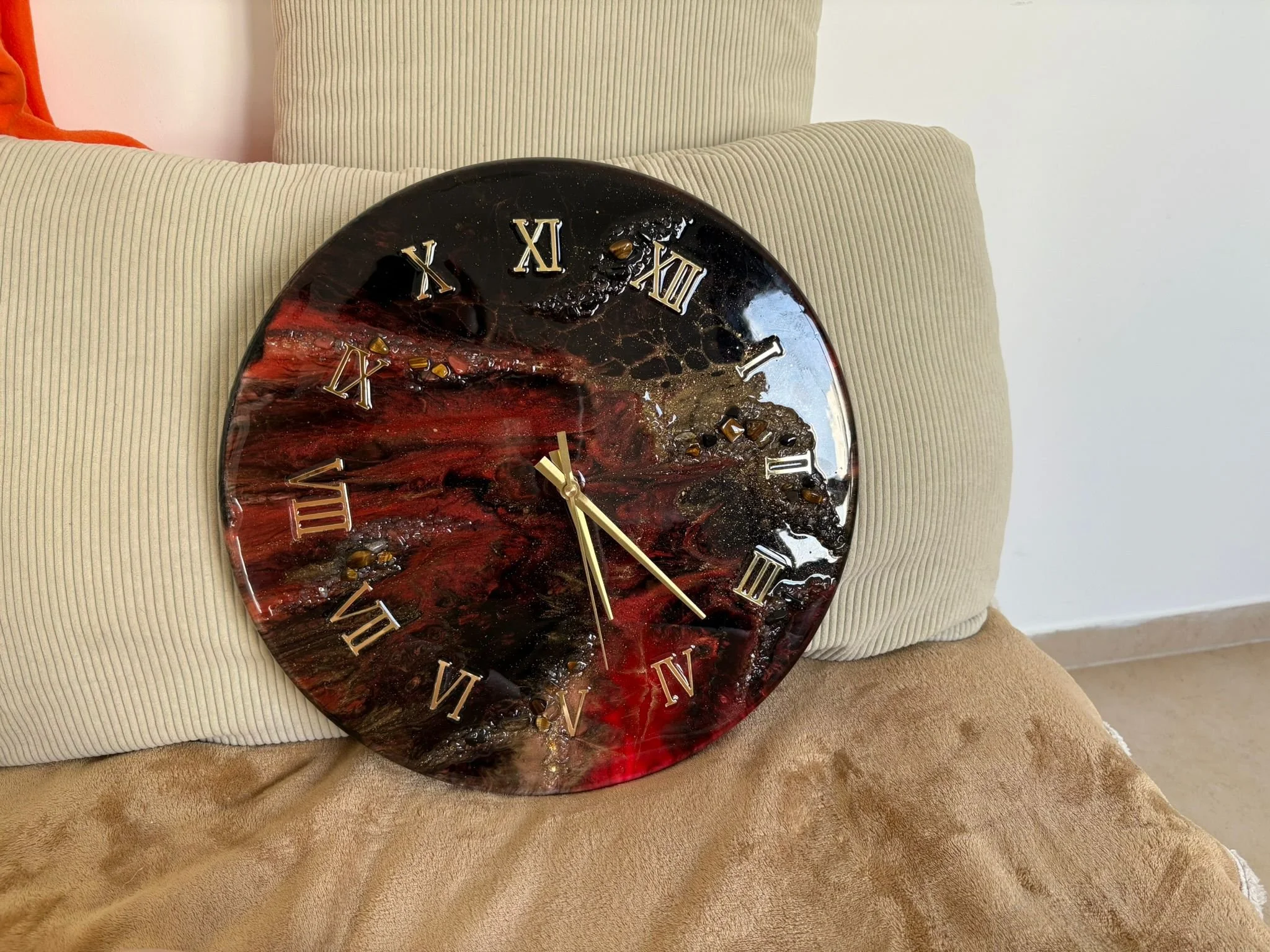 A round wall clock with a red and black marbled face and silver Roman numerals, resting on a beige couch cushion with beige pillows.