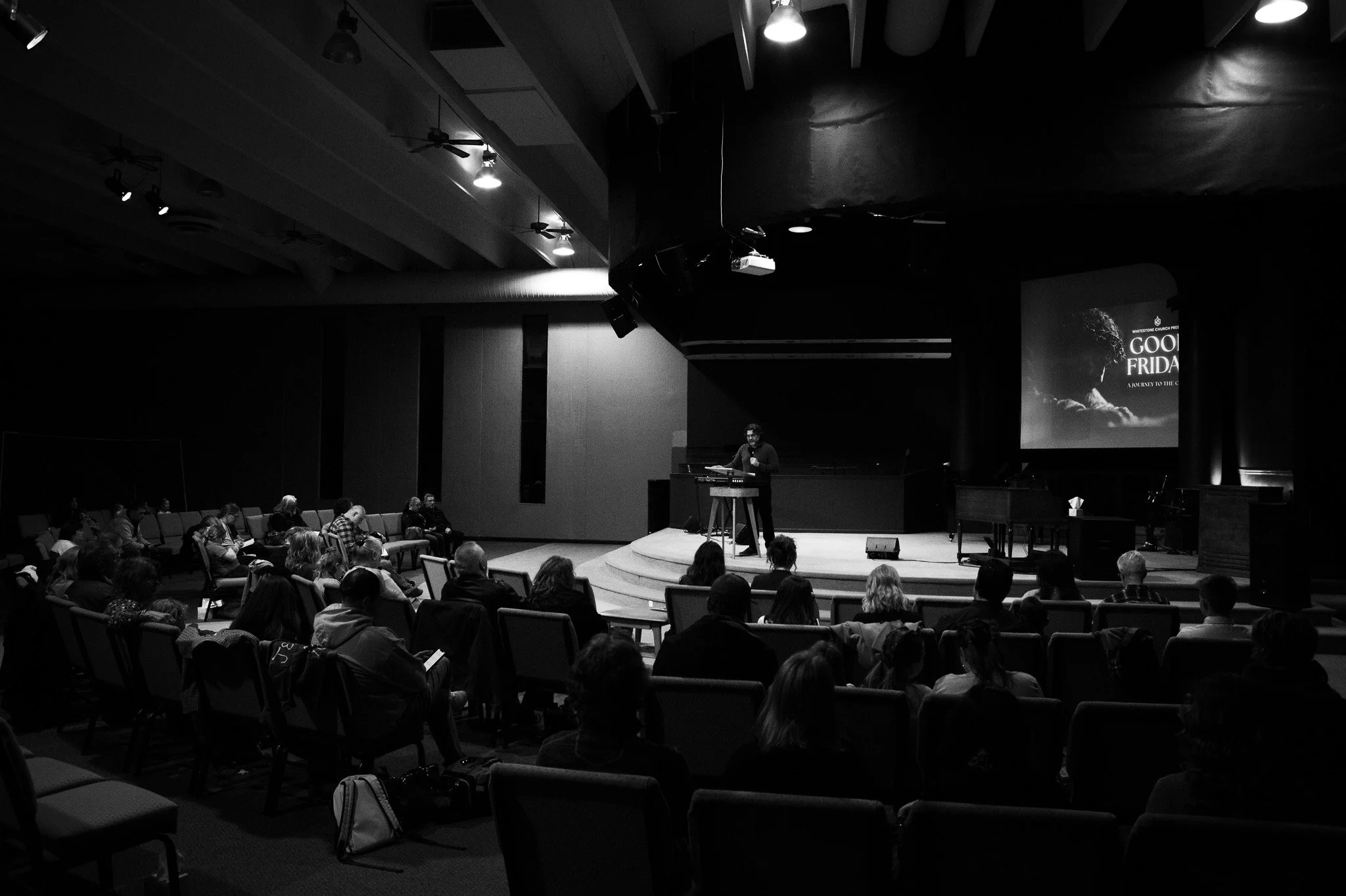 A man on stage giving a presentation to an audience at a conference or event. The stage has a large screen displaying a slide with the text "COOL FRIDAY" and a subtitle "A JOURNEY TO THE C...". The room is dimly lit, with spotlights on the stage and a few people in the audience taking notes or listening.