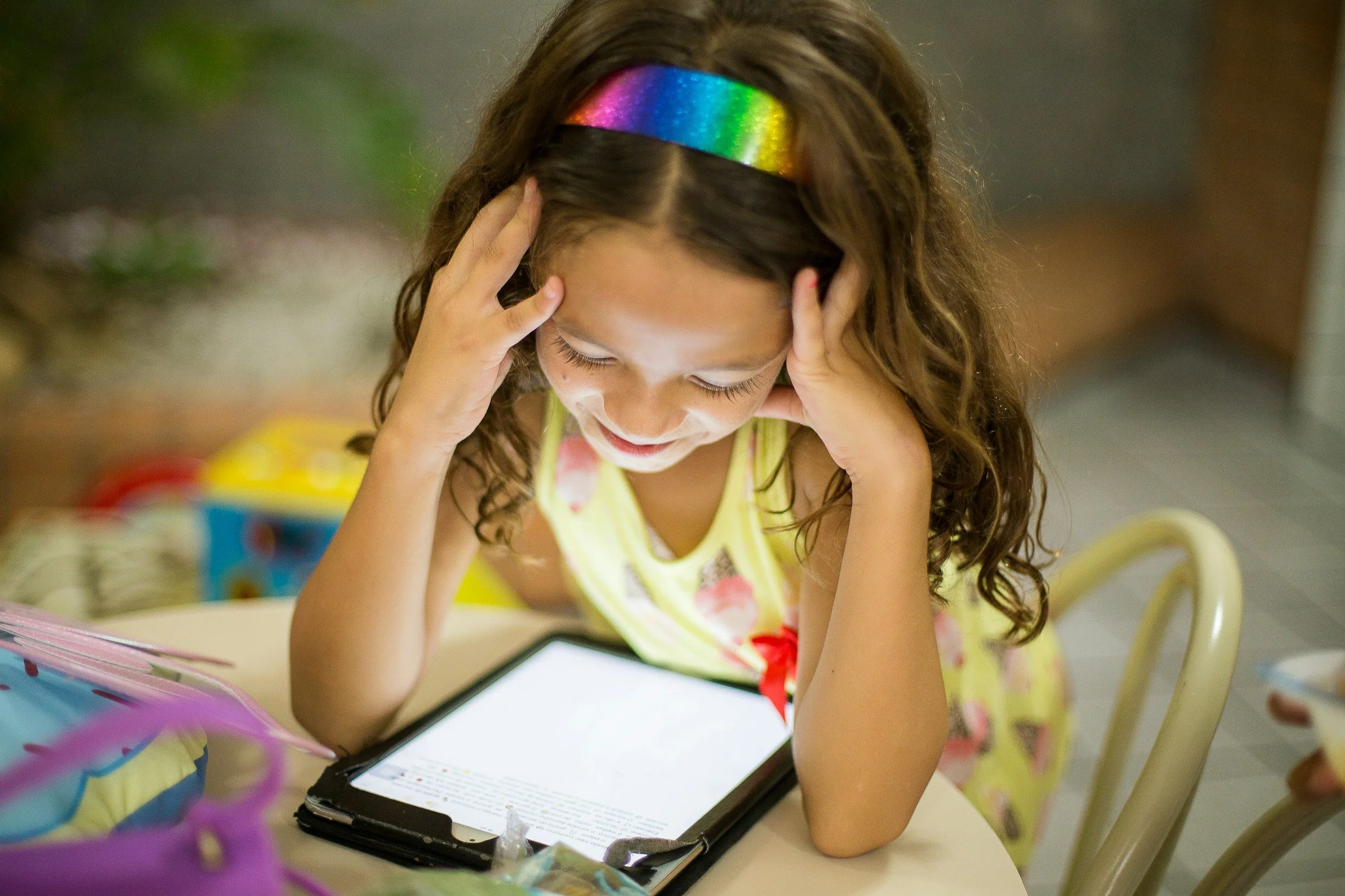 A young girl with curly hair and a rainbow-colored headband sits at a table, looking at a tablet screen with her hands on her head, and appears to be concentrating or stressed.