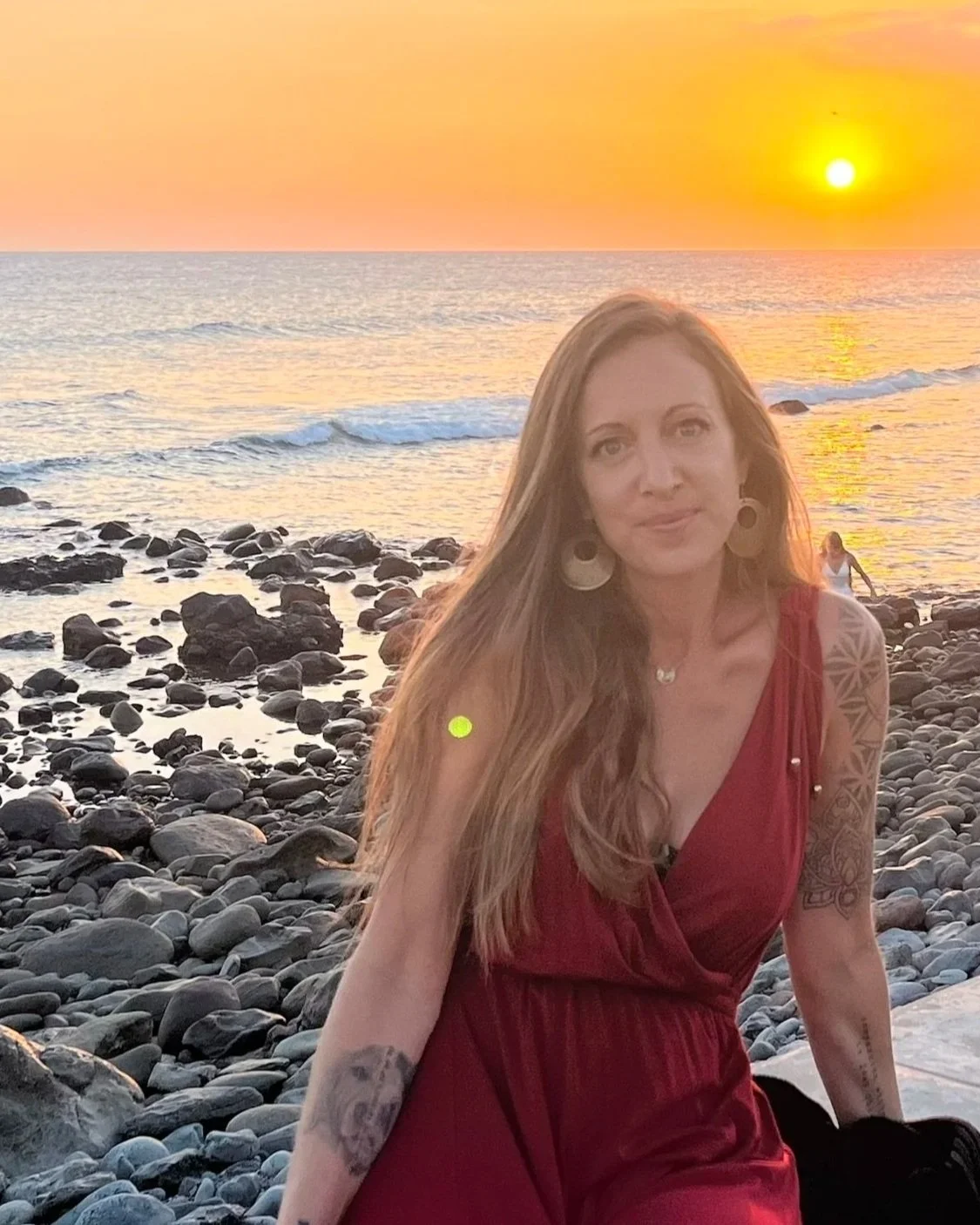A woman with long hair in a red dress sitting on a rocky beach at sunset.