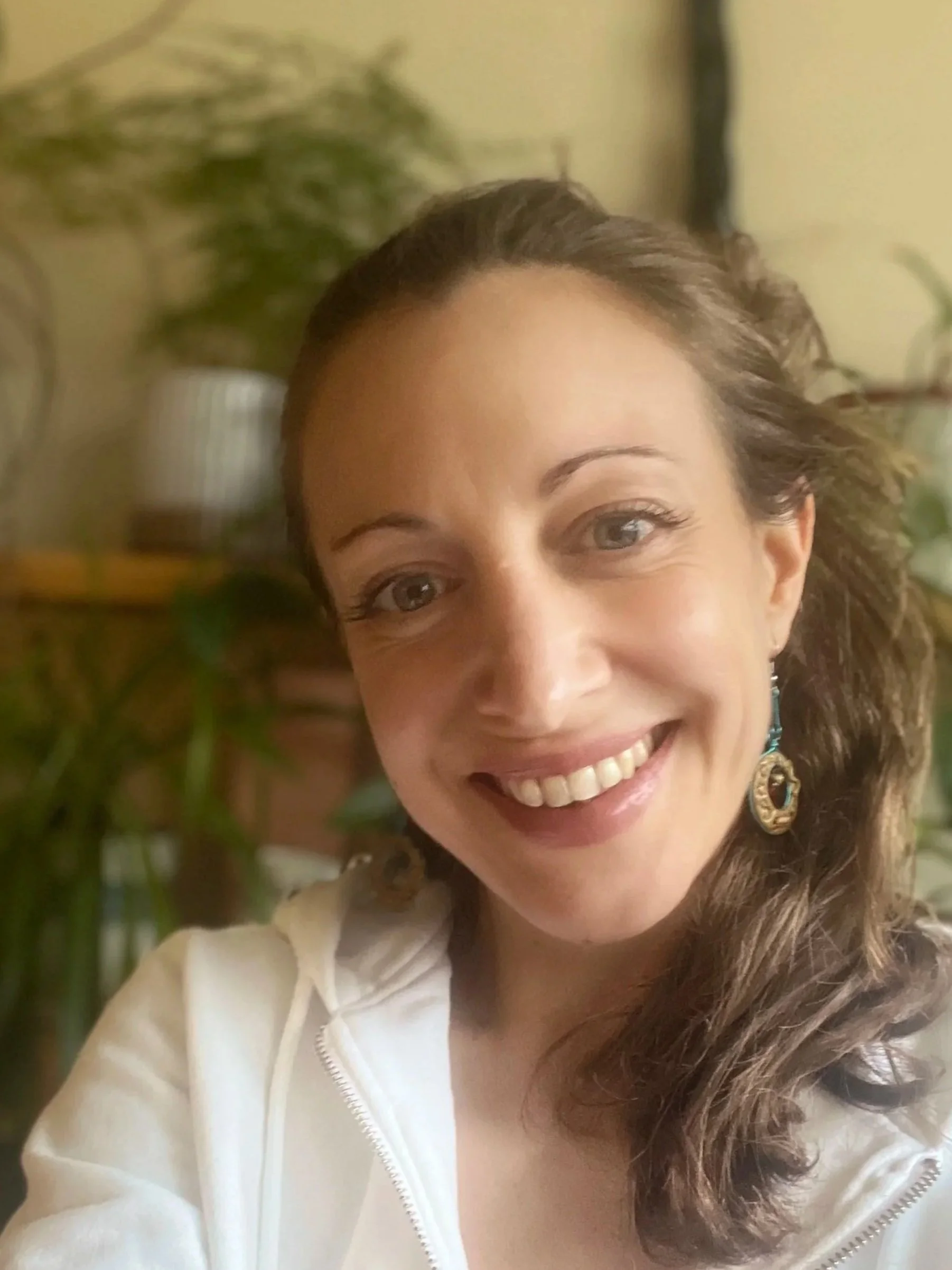 Close-up of smiling woman with curly brown hair, wearing large earrings, indoors with plants in the background.