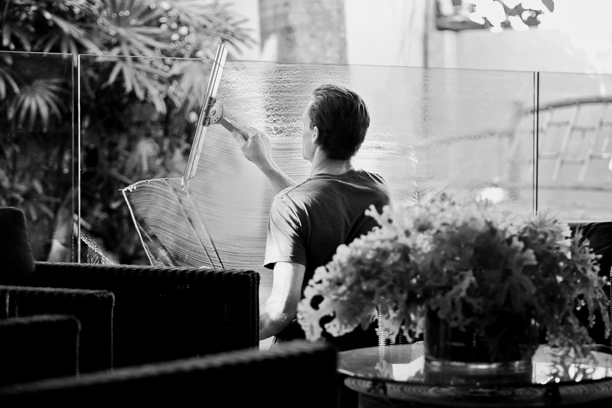 A person cleaning a large glass window or partition with a squeegee, seated indoors near some plants and a vase of flowers.