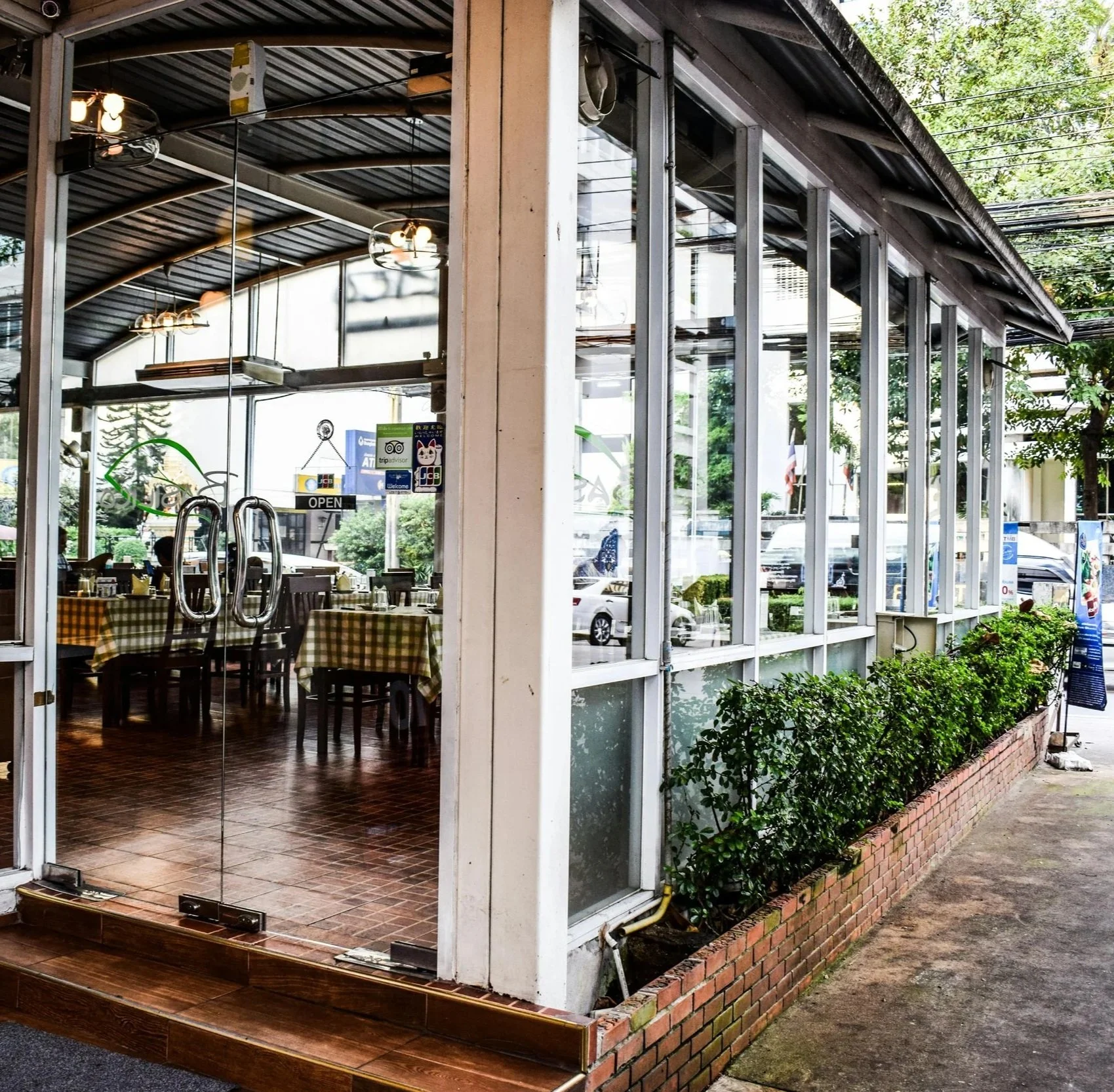 View of the exterior of a restaurant with glass walls and a garden bed with bushes outside. The restaurant's interior, visible through the glass, shows tables with checkered tablecloths and hanging pendant lights.