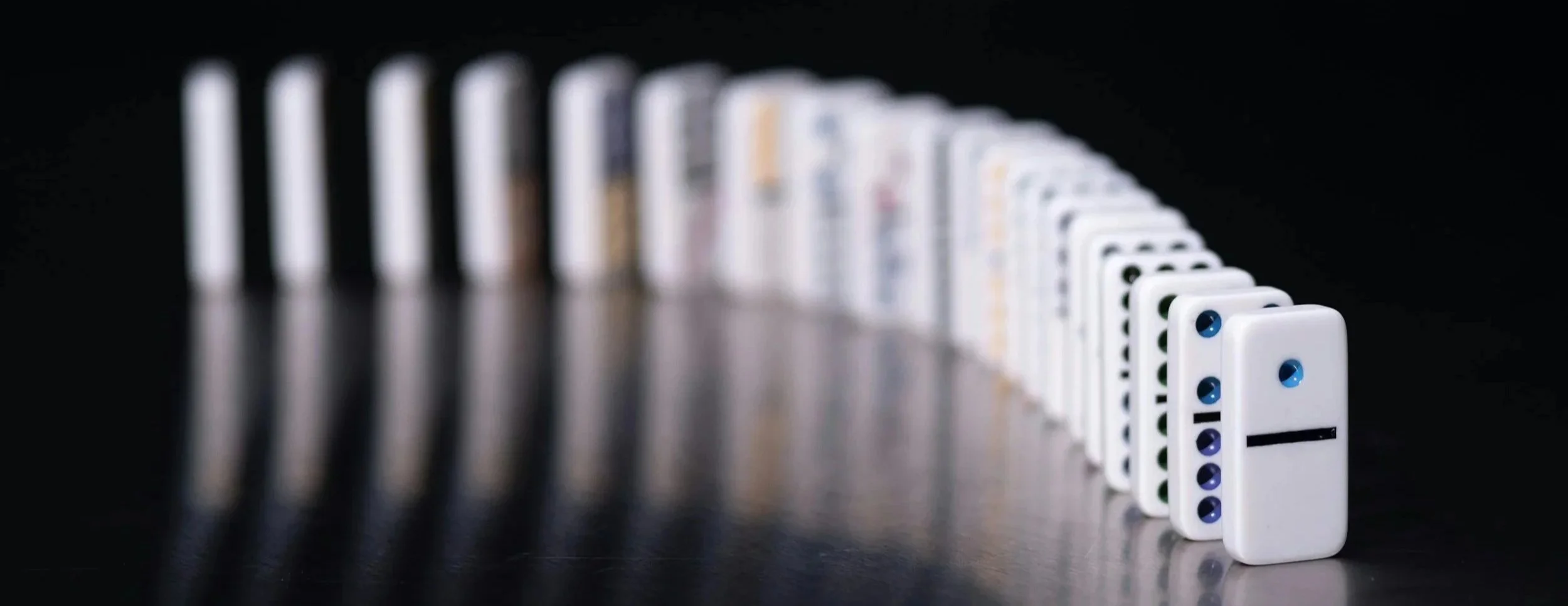 Row of dominoes falling in sequence on a reflective surface against a black background.