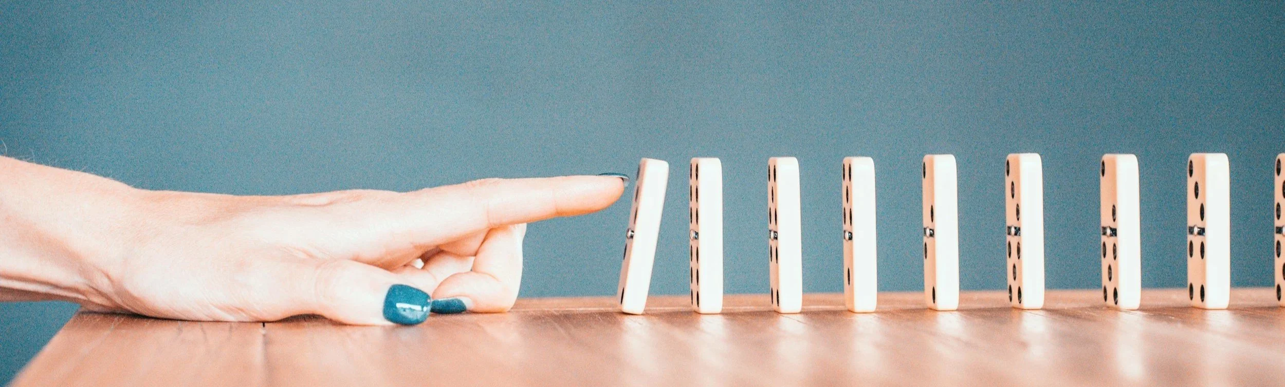 Hand with blue nails stopping a row of falling dominoes on a wooden surface against a blue background.