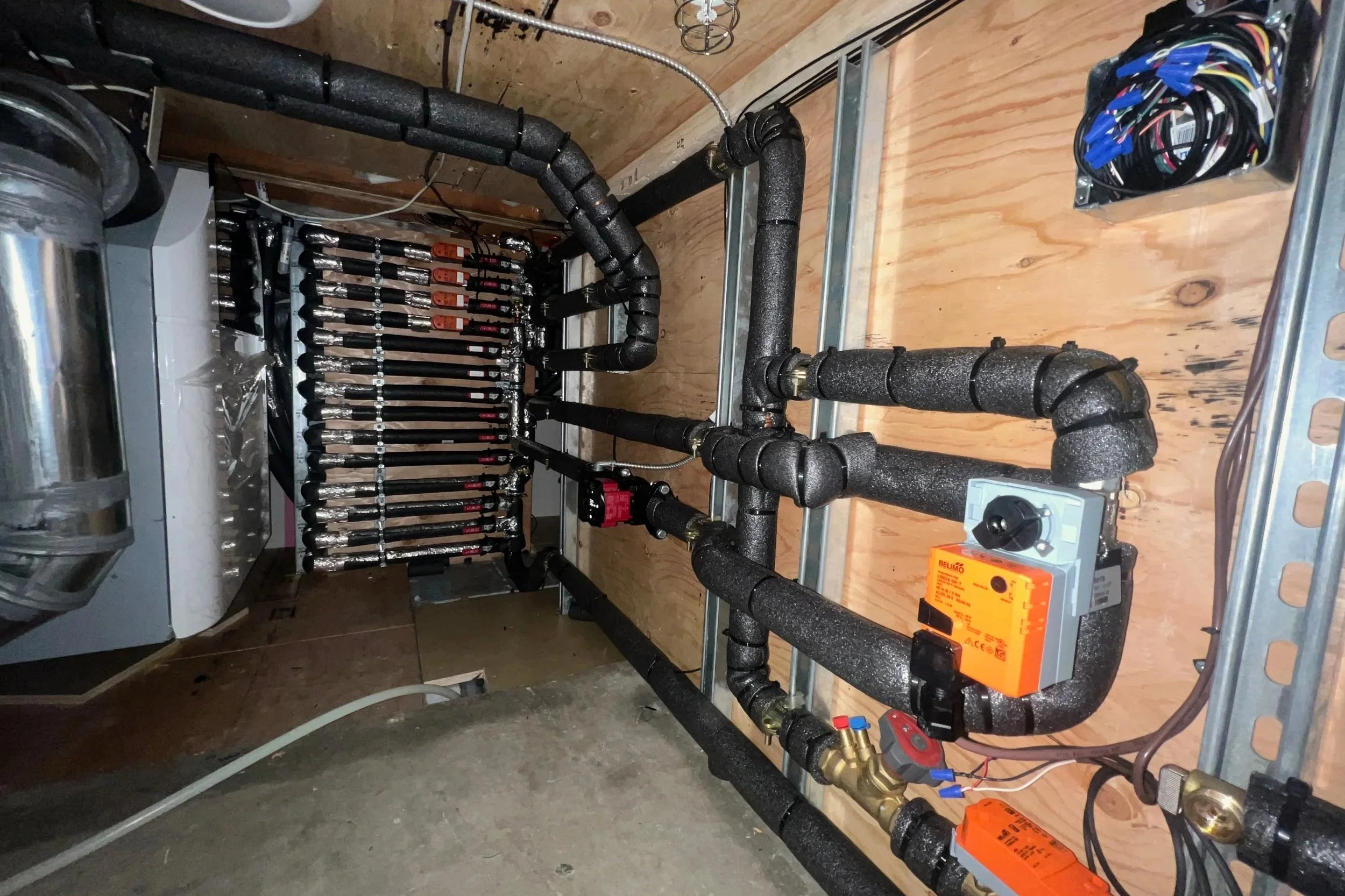 Mechanical room with black insulated pipes, flow control valve, and electrical wiring, mounted on a wooden wall.