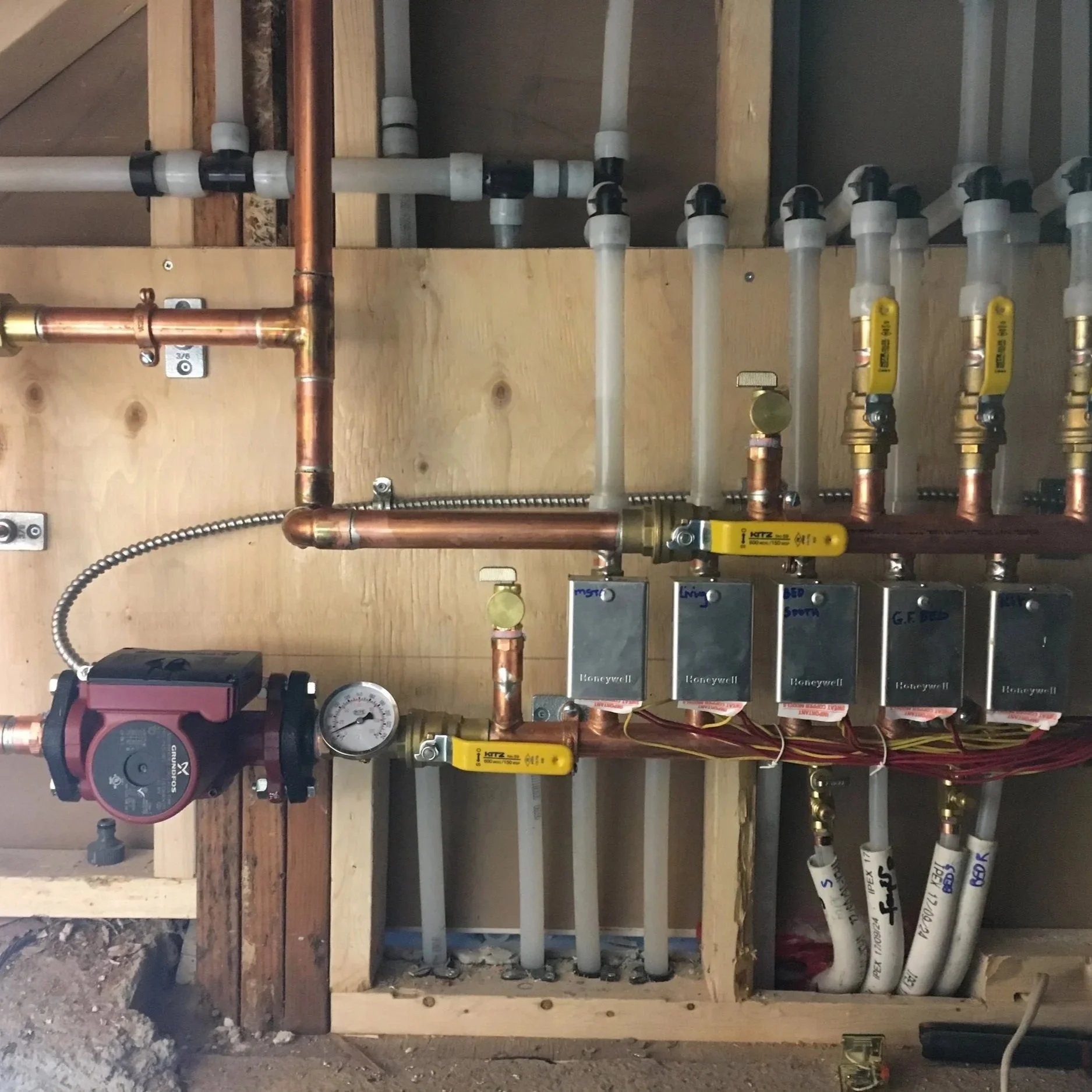 A plumbing system with copper and plastic pipes, valves, gauges, and electrical components installed inside a wooden wall frame.