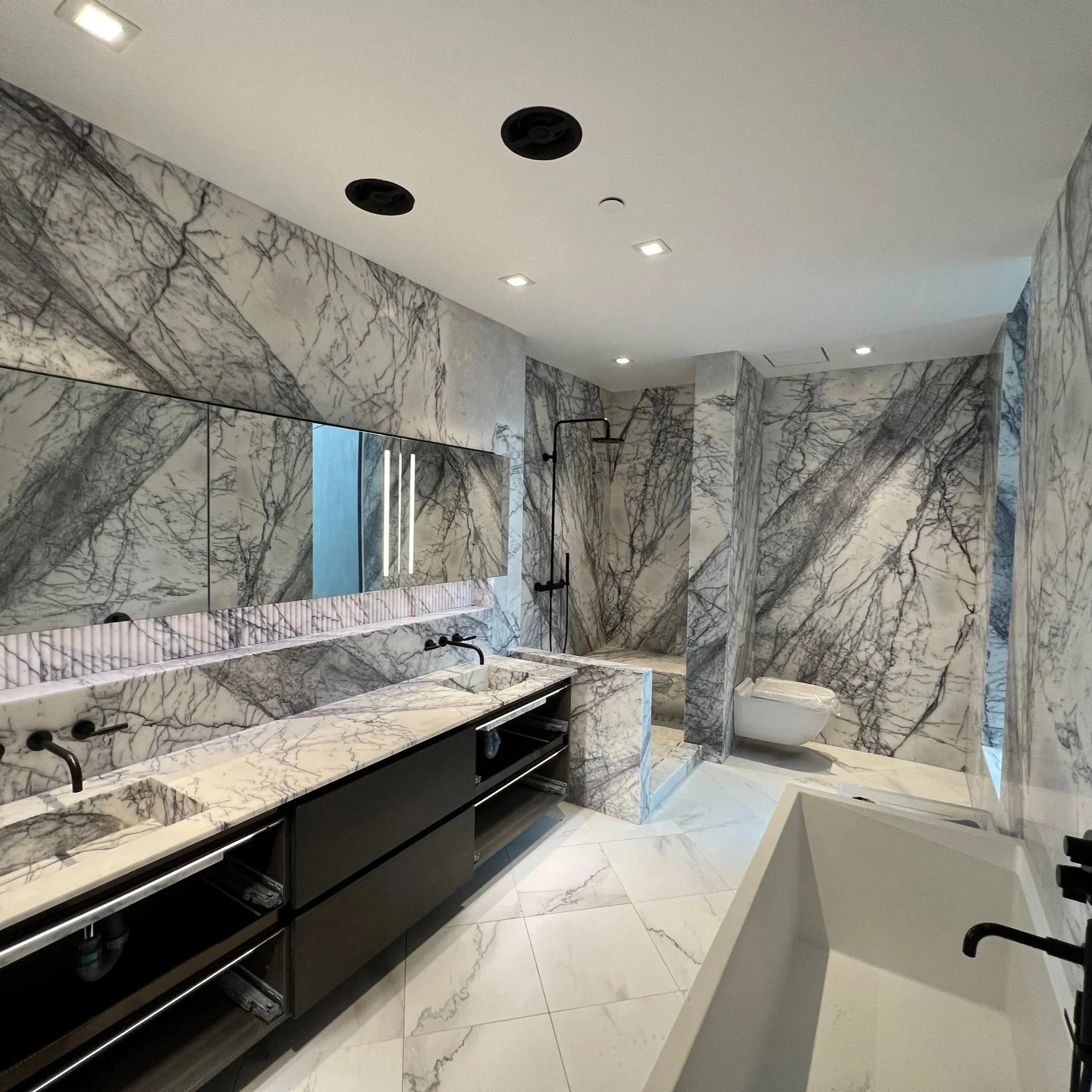 Modern bathroom with marble walls and floors, including a double sink vanity, a walk-in shower, a toilet, and a bathtub.