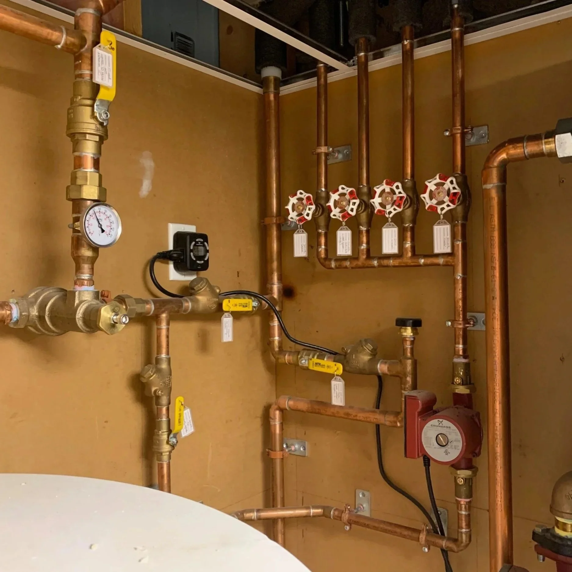 A plumbing installation with copper pipes, valves, a pressure gauge, and a red circulator pump mounted on a wall.