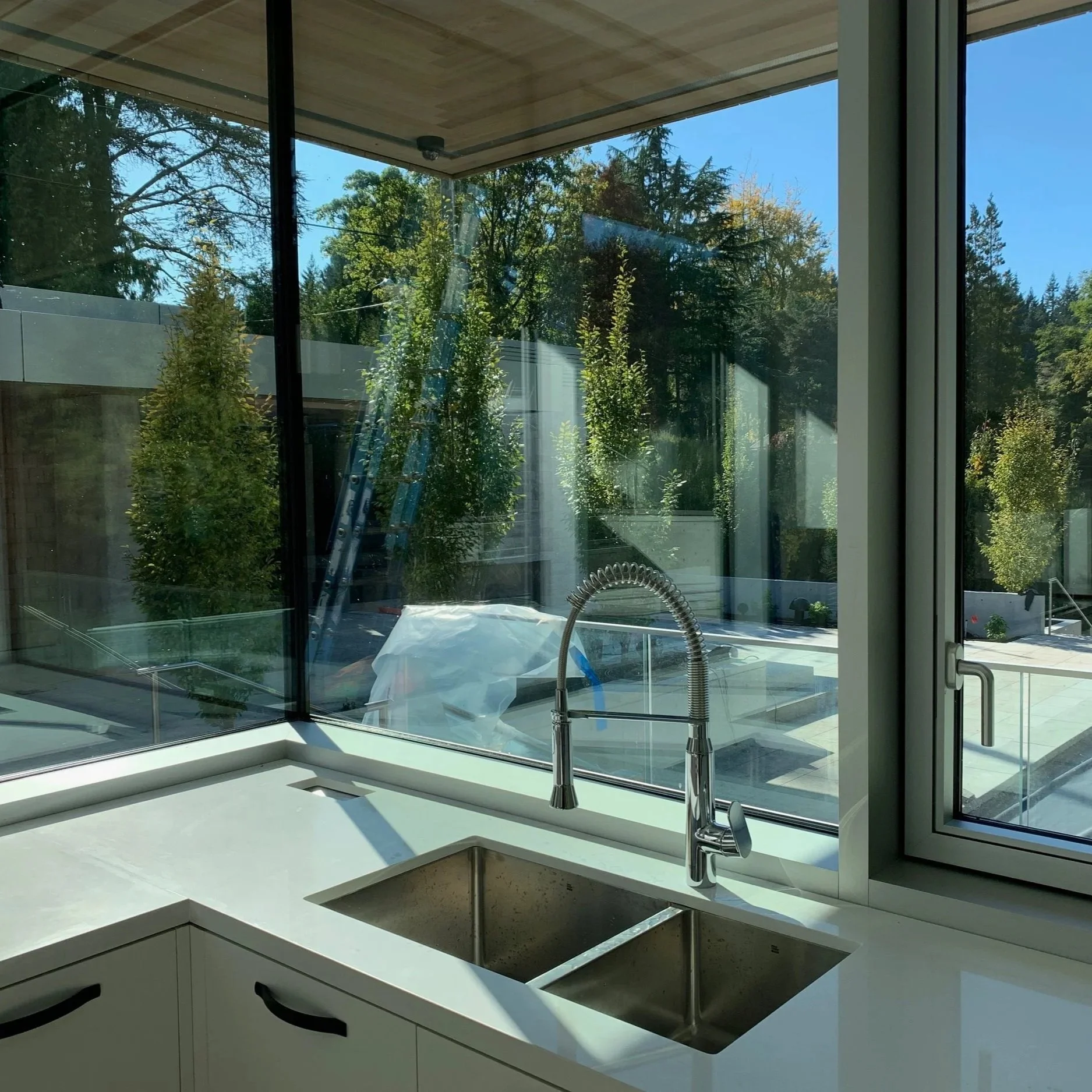 Kitchen sink with a modern chrome faucet inside a room with large windows overlooking a backyard with trees and outdoor furniture.