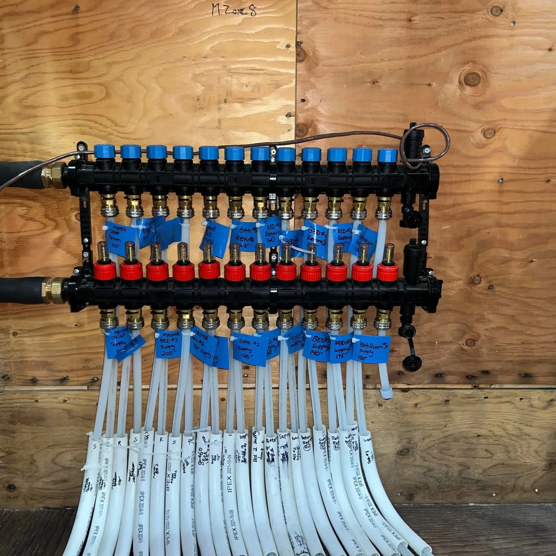 A plumbing manifold with multiple blue and red valves, connected to white pipes labeled for various rooms and purposes, mounted on a wooden wall.