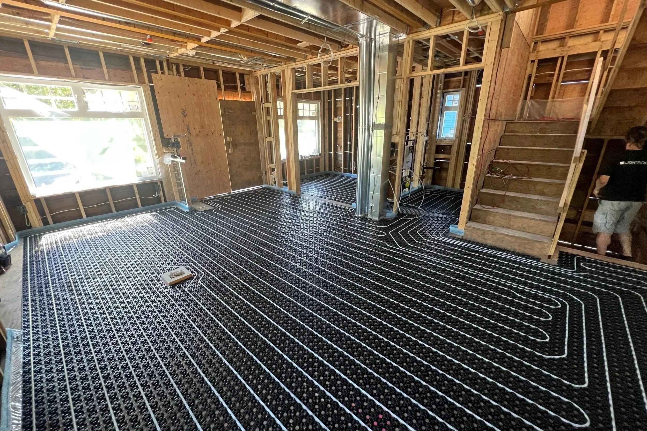Interior of a house under construction with radiant floor heating system installed, showing black panels with white tubing and exposed wooden framing.