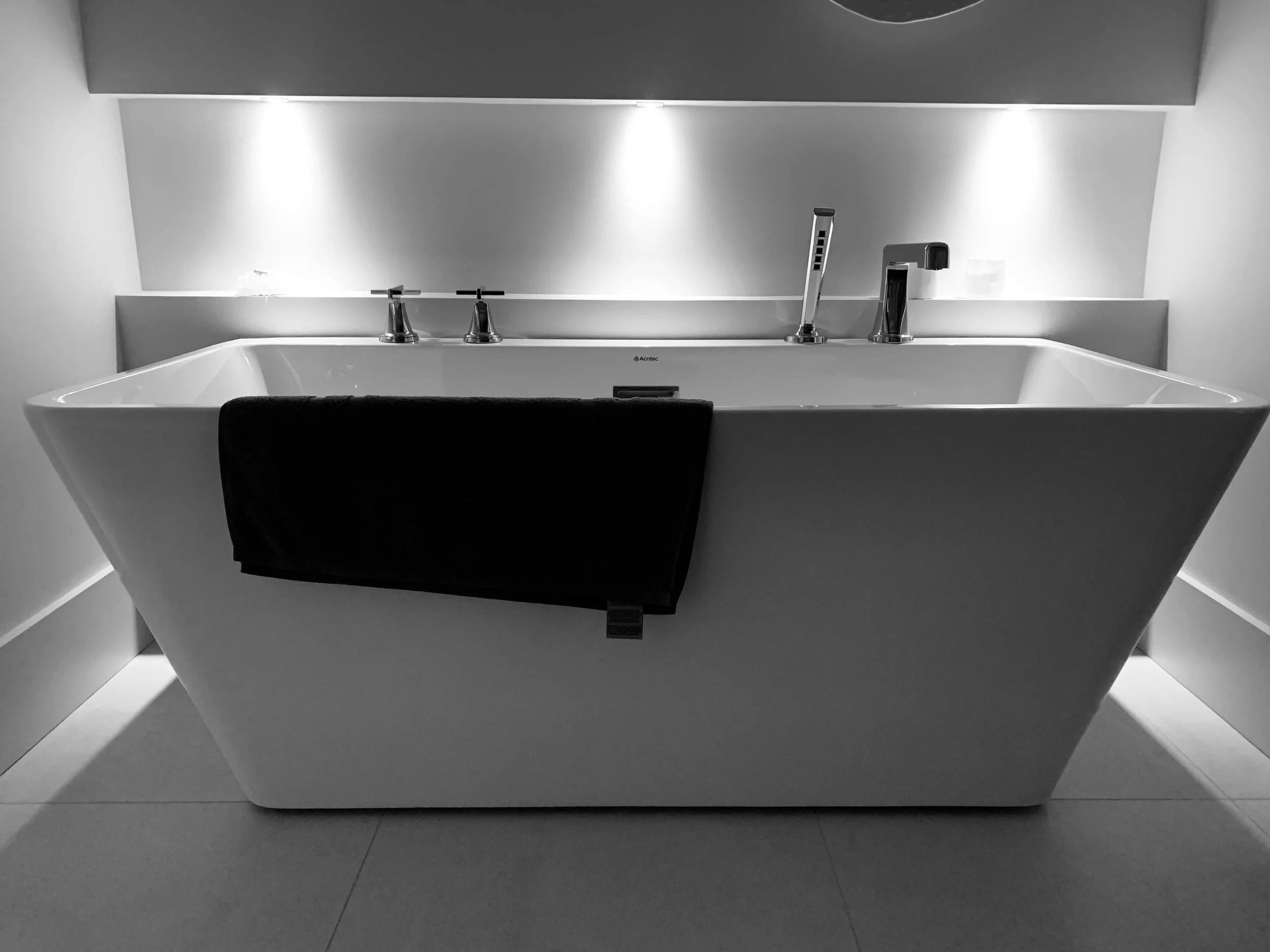 A modern white bathtub with a black towel hanging over the front, located in a minimalist bathroom with a mirrored wall and contemporary fixtures.