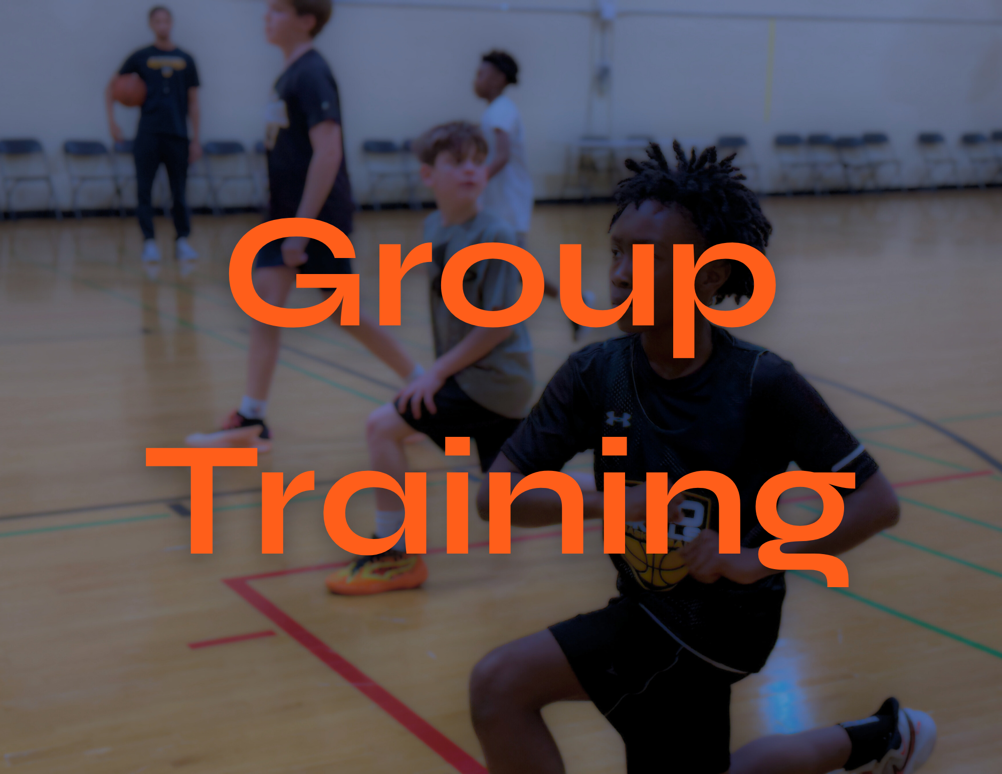 Children participating in a basketball training session indoors, with one child kneeling on the floor holding a basketball, and others standing behind, in a gymnasium.