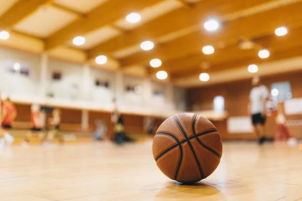 A basketball on an indoor basketball court with players and bright ceiling lights in the background.