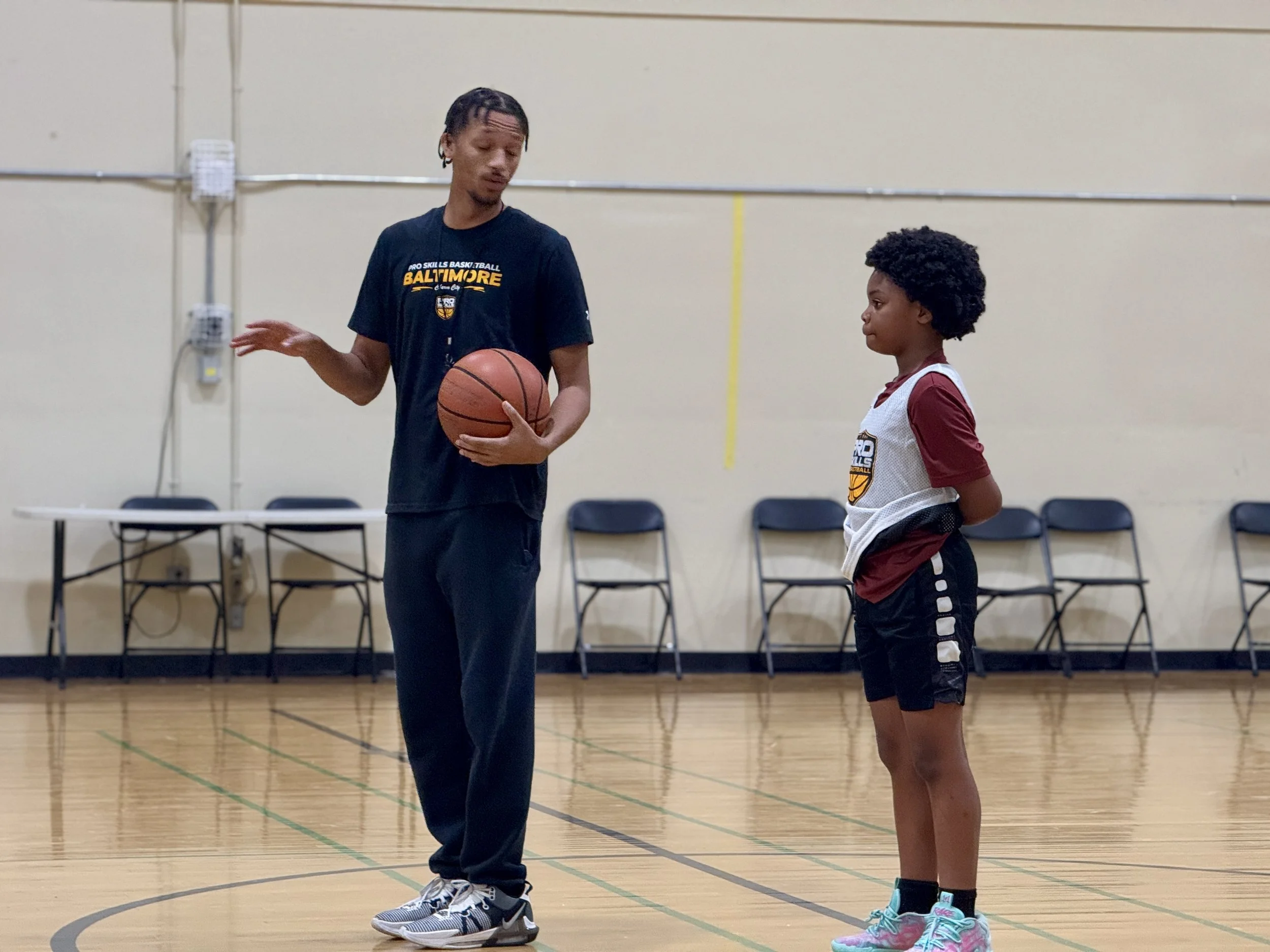 A young girl standing on a basketball court, listening to a male coach holding a basketball.
