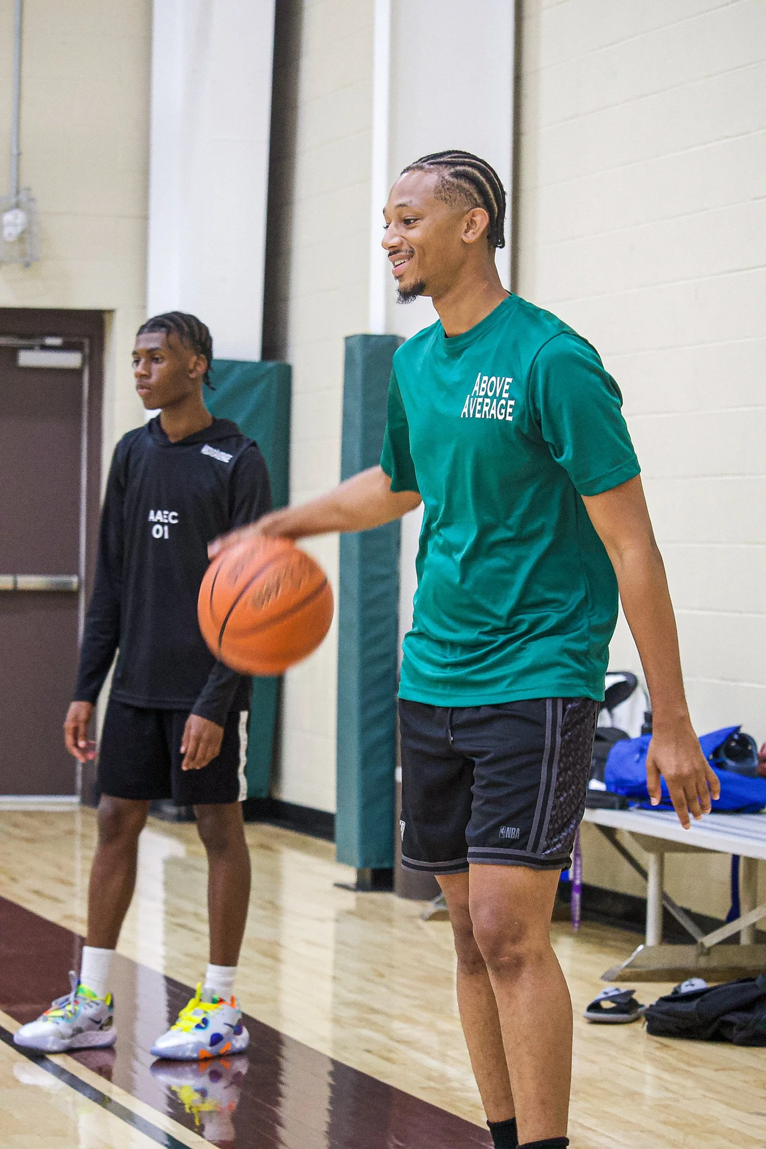 Two young men in a gymnasium, one holding a basketball and smiling, the other standing nearby, both in casual athletic clothing.