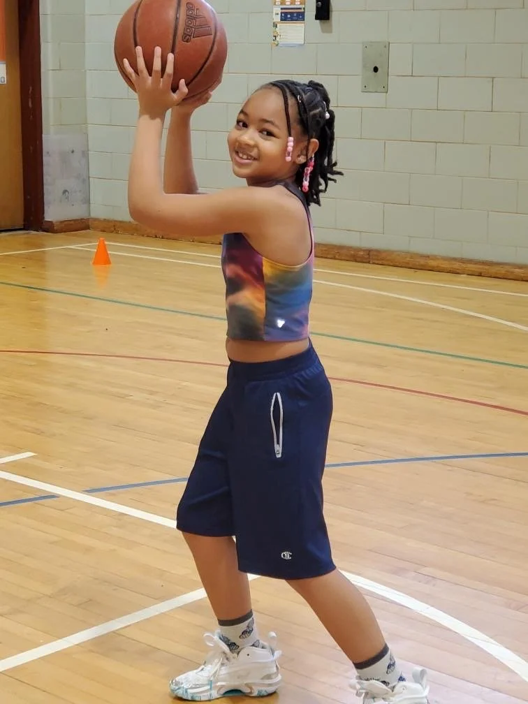 Young girl in a gym, holding a basketball above her head, smiling. She has dreadlocks with beads, wearing a colorful sleeveless top, navy shorts, white sneakers, and patterned socks.