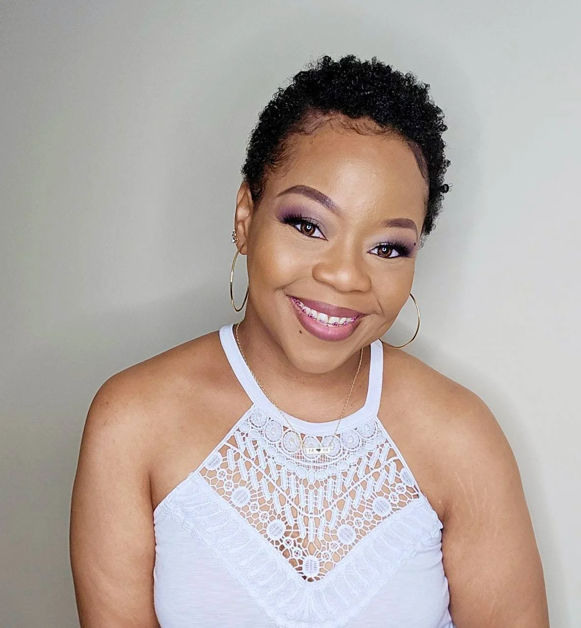 A smiling woman with short, curly black hair, wearing a white lace sleeveless top, gold hoop earrings, and a delicate gold necklace, standing against a plain light-colored background.