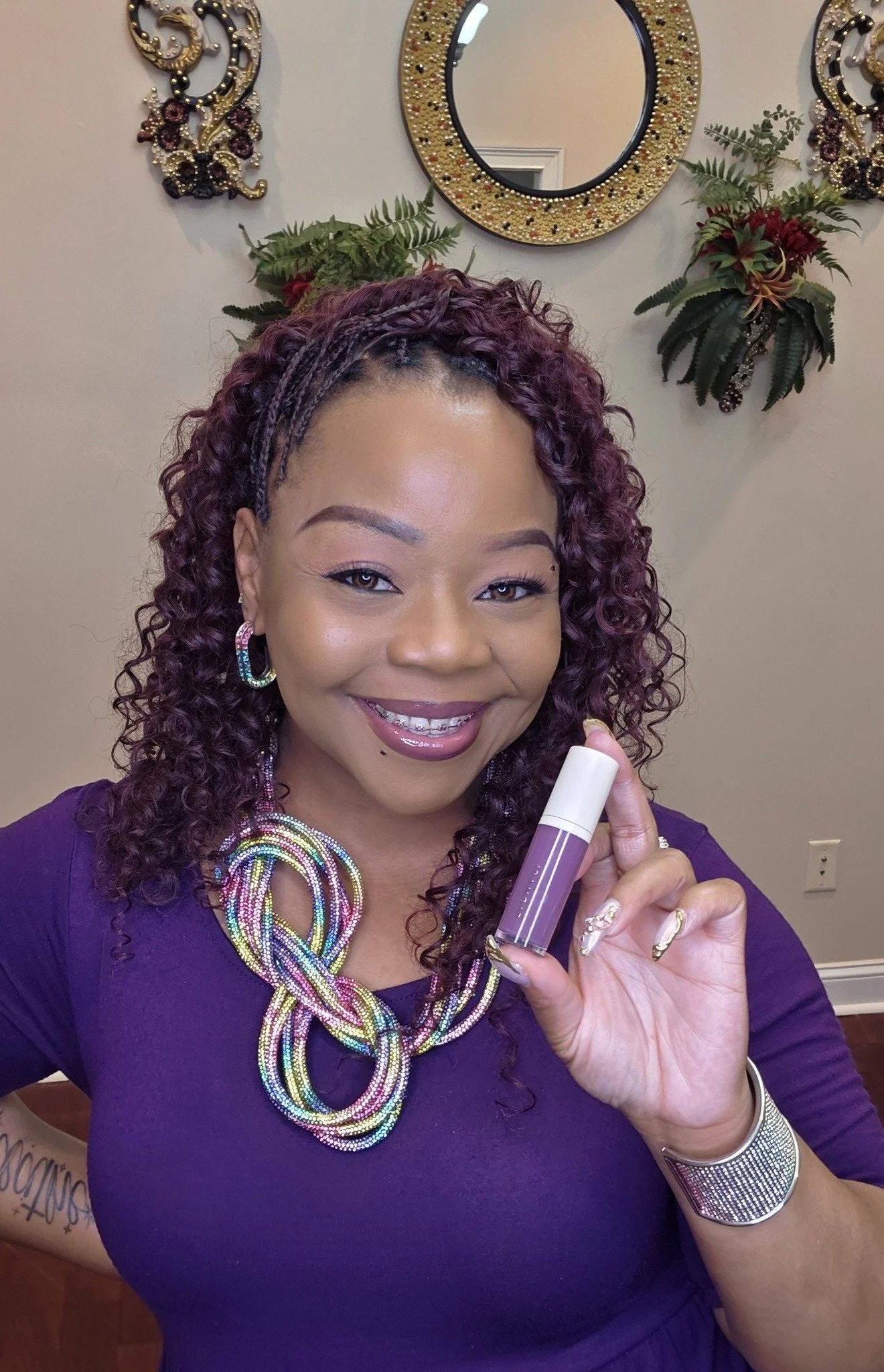 A woman with curly purple hair smiling while holding a purple lipstick tube. She is wearing colorful jewelry and a purple top, standing in a room with decorative wall art, mirror, and plants in the background.