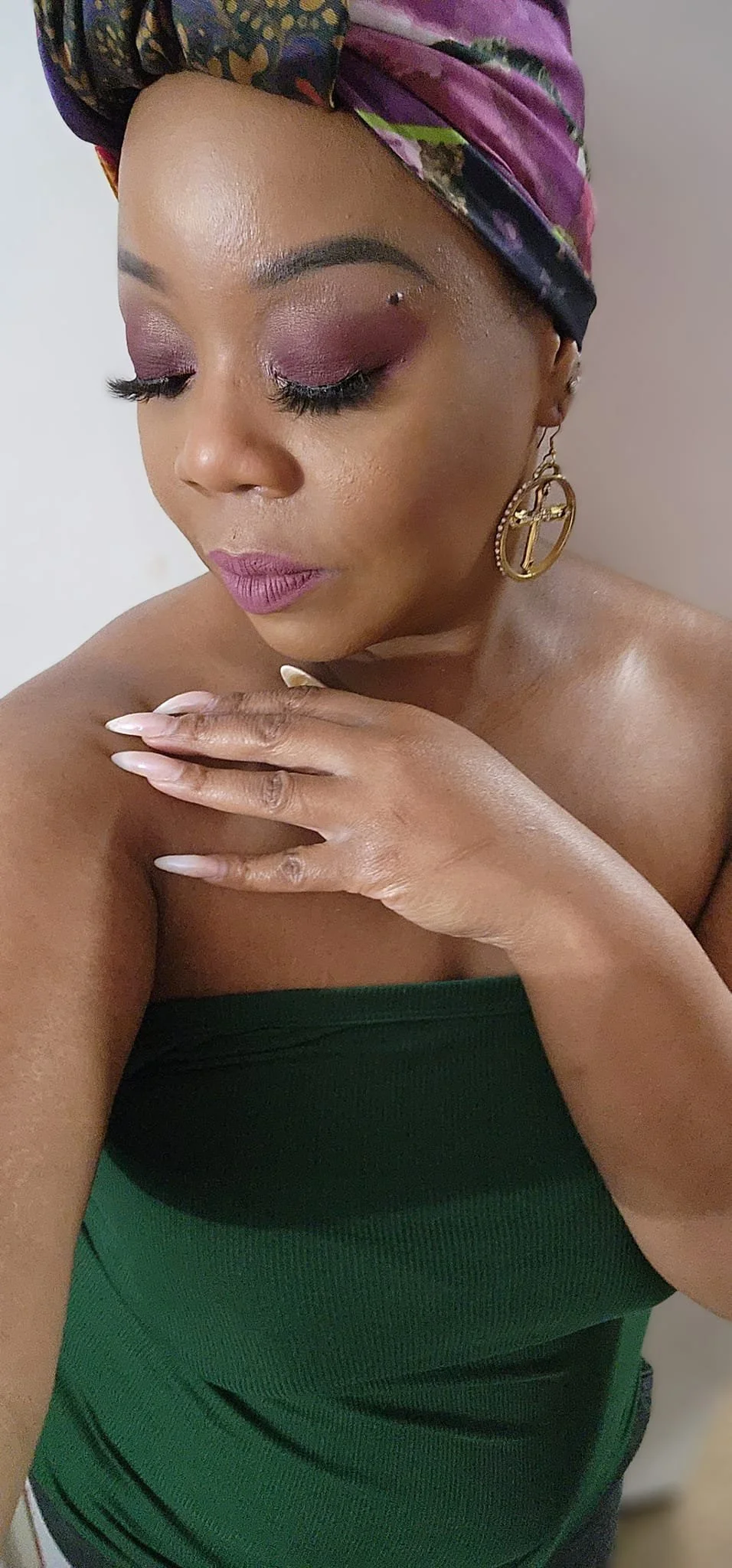 A woman with dark skin, makeup, and a colorful headscarf, wearing a strapless green top and large circular earrings, posing with her hand on her shoulder and eyes closed.