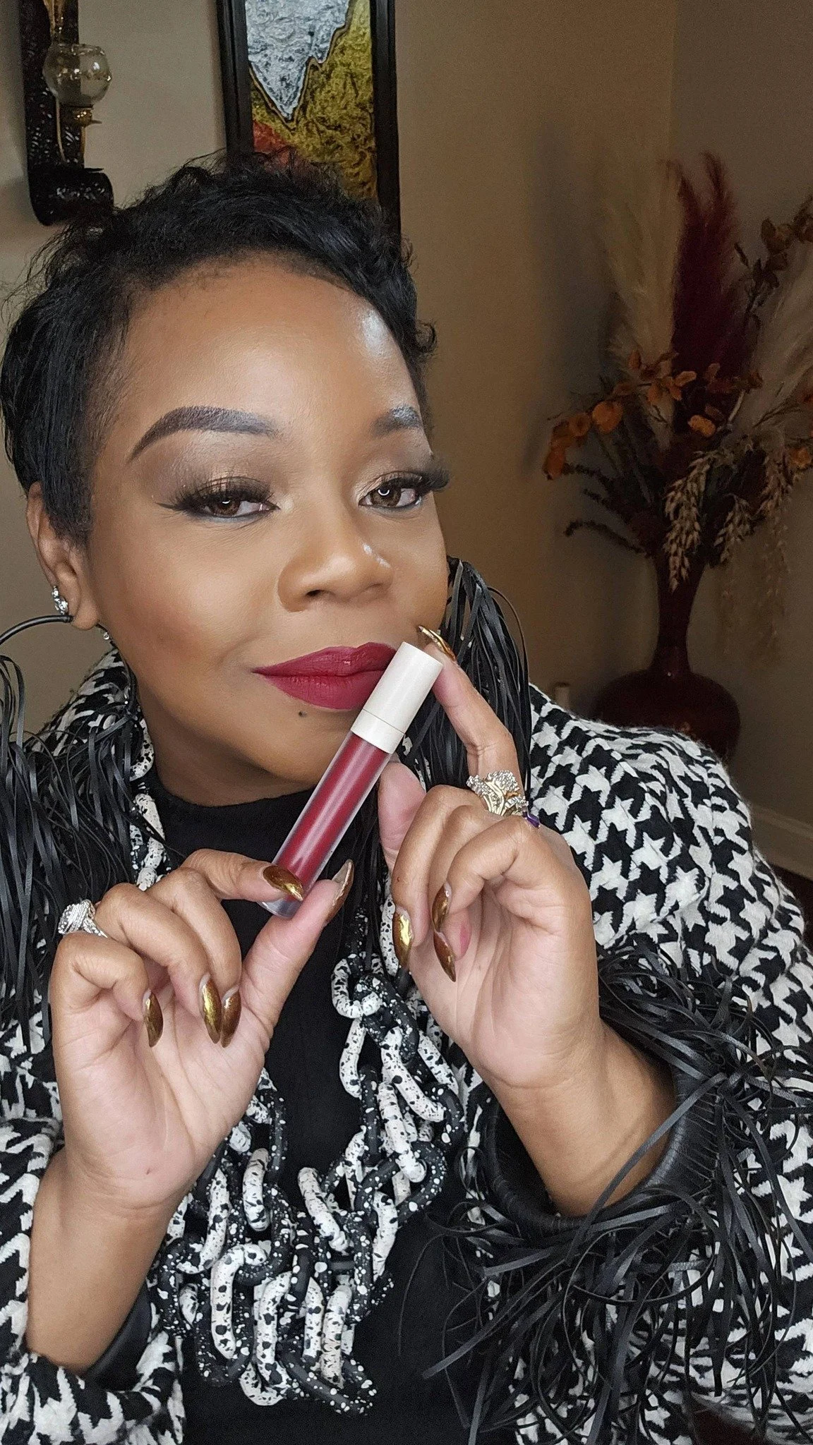 A woman with short black hair and makeup holding a lip gloss tube, wearing a black and white patterned outfit with decorative rings and gold metallic nail polish, sitting in a room with art and decorative plants.