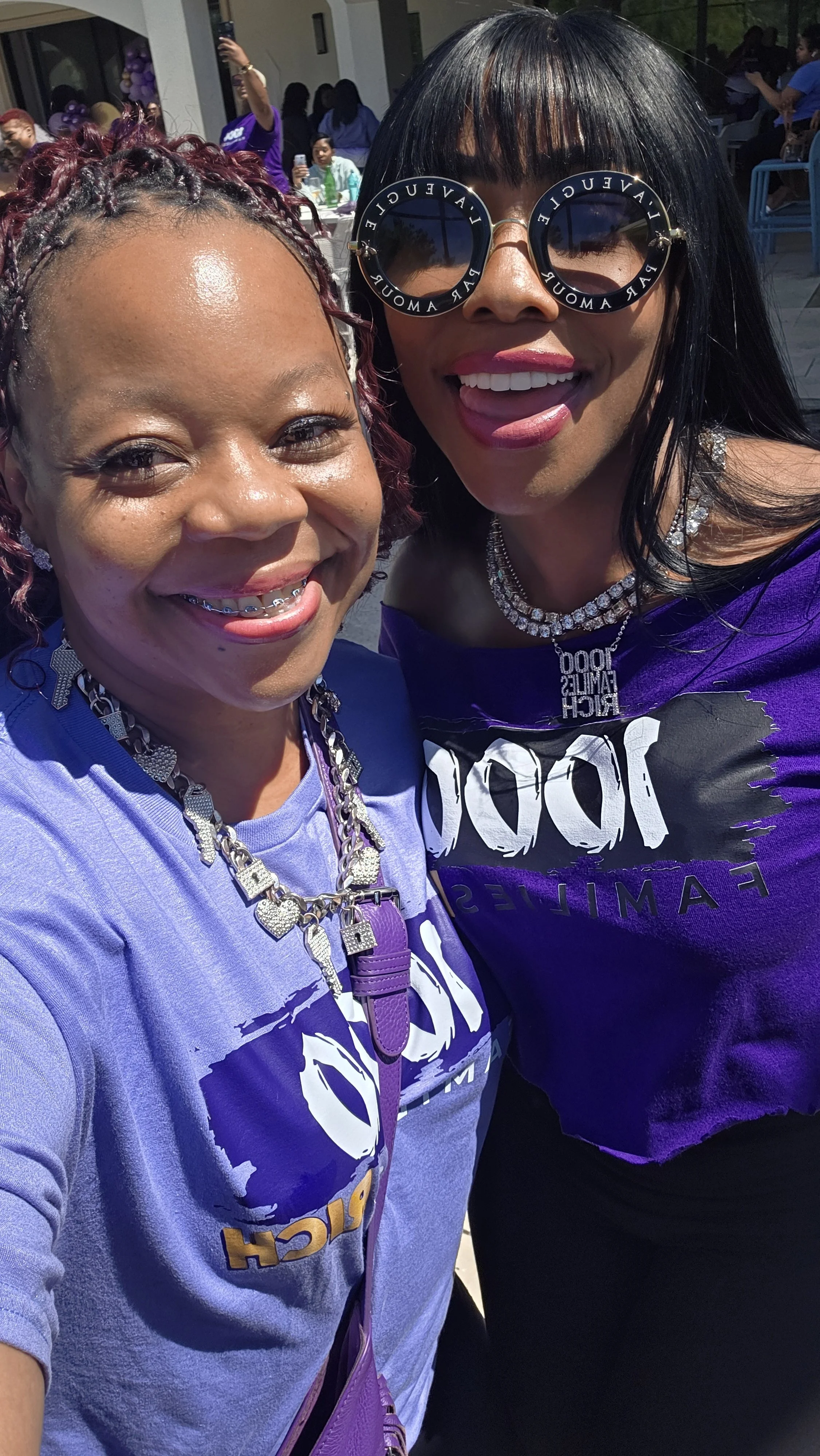 Two women smiling for a selfie outdoors, wearing purple shirts, with several people and balloons in the background.