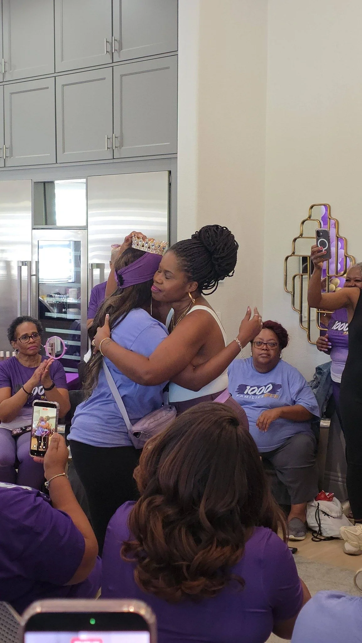 Two women hugging warmly, one wearing a crown and purple headband, surrounded by seated women in purple shirts capturing the moment.