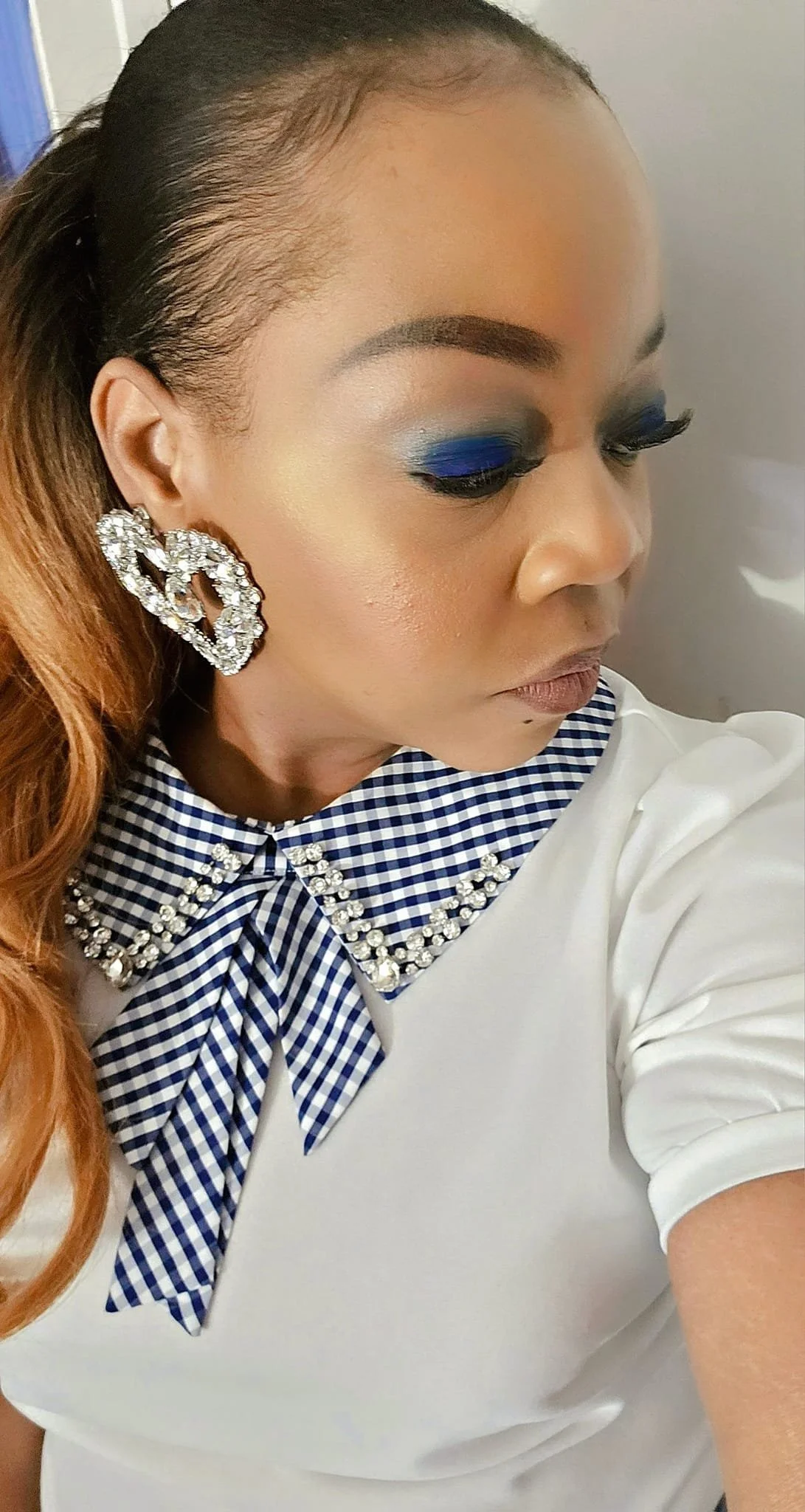 Close-up of a woman with styled hair, bold eye makeup with blue eyeshadow, large rhinestone earrings, and a white top with a plaid bow and embellished collar.